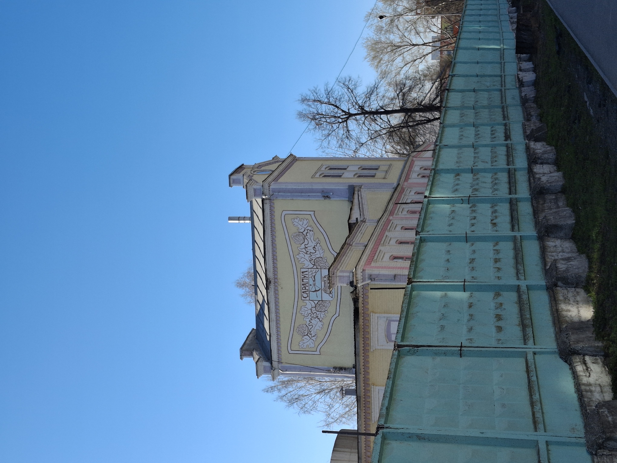 A bright daytime scene captures a distinctive building alongside a long barrier under a clear blue sky. The building, oriented horizontally across the mid-ground of the image (when correctly rotated), features a light yellow facade adorned with elaborate light-colored relief or painted murals. A prominent decorative panel on the building displays the Cyrillic text "САРАПУЛ" (SARAPUL), surrounded by stylized foliage, possibly oak leaves and acorns, and a small emblem resembling a boat on water. The architecture includes multiple stories with various window shapes and light pinkish-red accents on some window frames or decorative elements.
In the foreground, running parallel to the building, is a substantial light turquoise/teal barrier or fence made of textured panels, possibly concrete, indicating a boundary or retaining wall. Beyond this barrier and to the right, patches of green grass and rugged concrete or stone walls are visible, suggesting a slope or embankment. A dark gray paved surface, likely a road or sidewalk, is present on the far right.
Above the building and against the vast, clear blue sky, several bare deciduous trees stand, their branches devoid of leaves, suggesting a season like late autumn, winter, or early spring. The bright lighting indicates a sunny day. No specific human activity or people are visible, presenting a static architectural and landscape view.