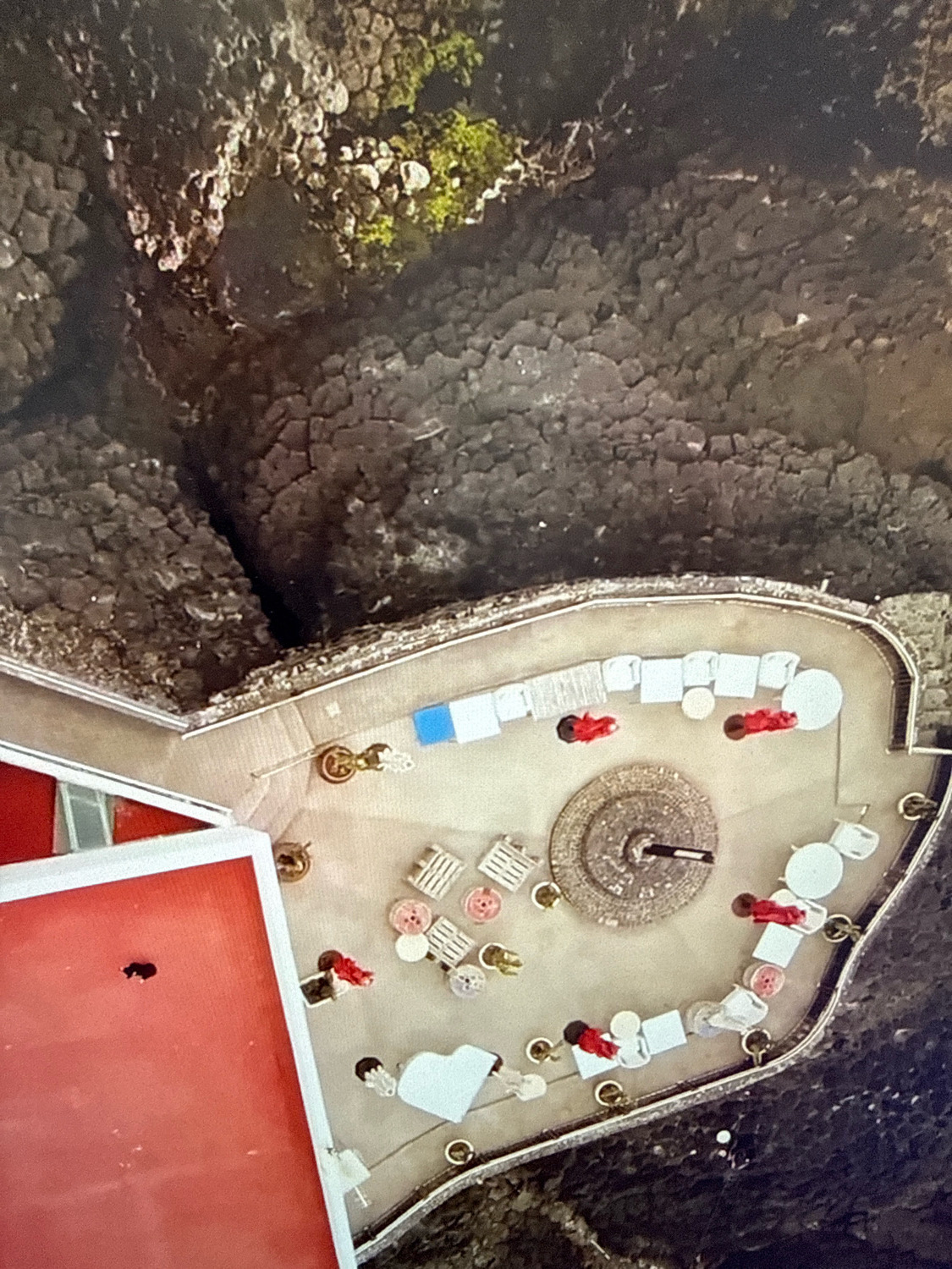 This is a high-angle aerial view of an outdoor seating area. The area is paved and features several white tables and chairs, as well as some lounge chairs. There are also several people present, wearing red clothing, seated at the tables and standing. In the center of the seating area is a circular stone structure. To the left, a red building with white trim is visible. The background is composed of dark, rocky terrain and some greenery. The location is the Cliffside Restaurant in Netanya, Israel.