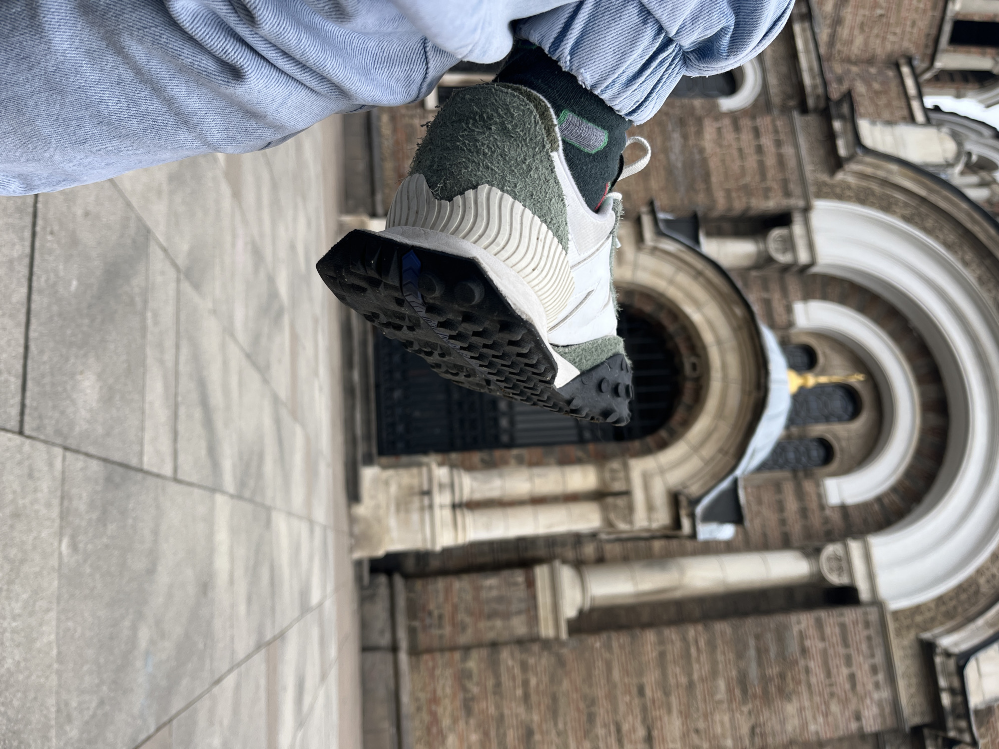A person wearing a light blue denim jacket and black socks stands in front of what appears to be the facade of the Saint Sofia Church in Sofia, Bulgaria. The focus is on the sole of their green, white, and black sneaker. The backdrop includes the church's architectural details, like its stone brickwork and arched windows. To the left, the pavement is visible, with stone brickwork running vertically to the bottom left.
