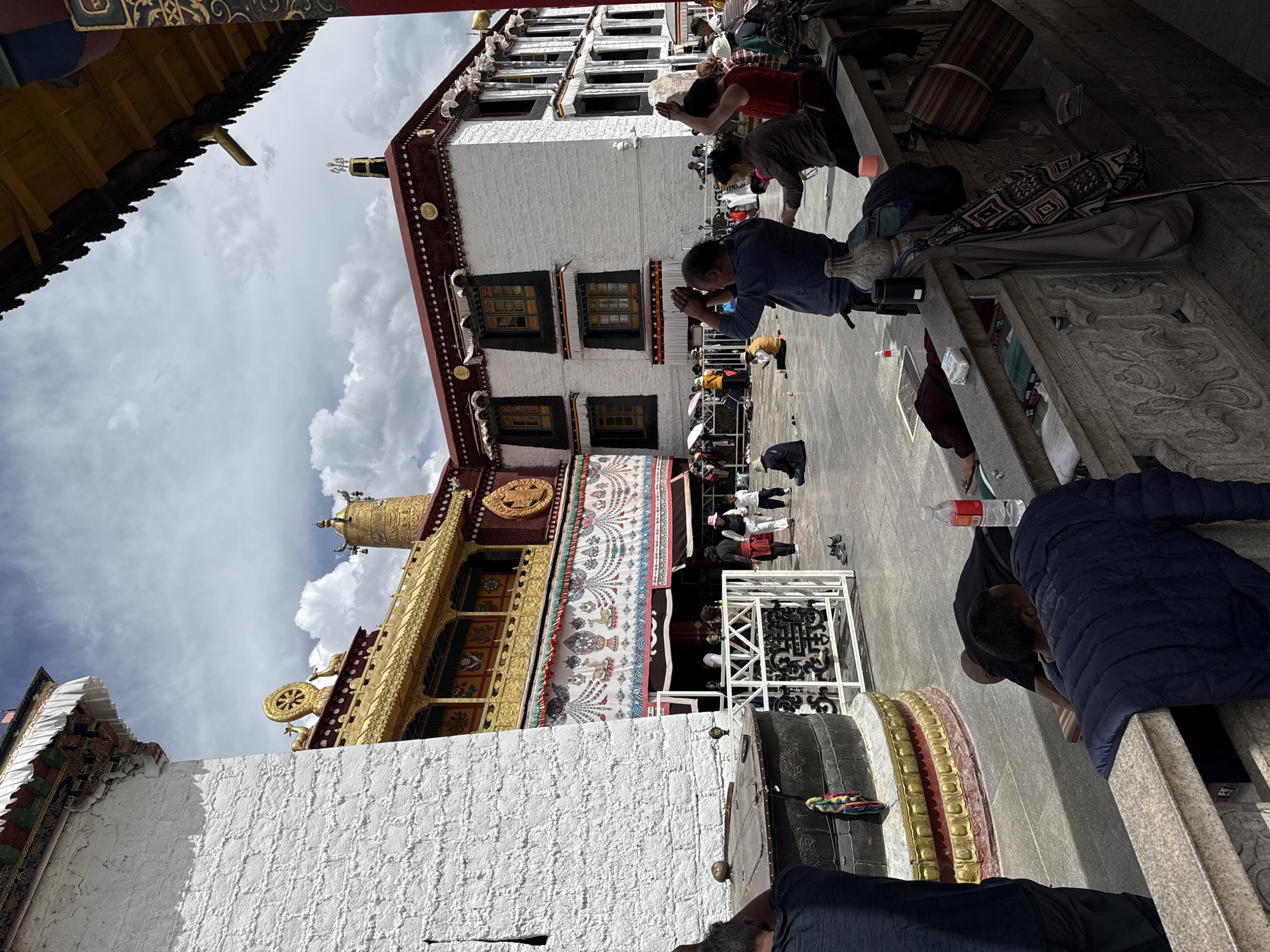 The image shows a view in front of the Jokhang Temple in Lhasa, Chengguan District, China. The temple buildings have a traditional Tibetan architectural style with white walls, red and gold trim, and ornate facades. Several people are present, some standing and praying with hands clasped, while others are kneeling or prostrating themselves in apparent acts of worship or devotion. Some people also are walking around and standing in the temple complex. The environment is an open courtyard or plaza, with stone steps and ornate carvings. The sky is partly cloudy.