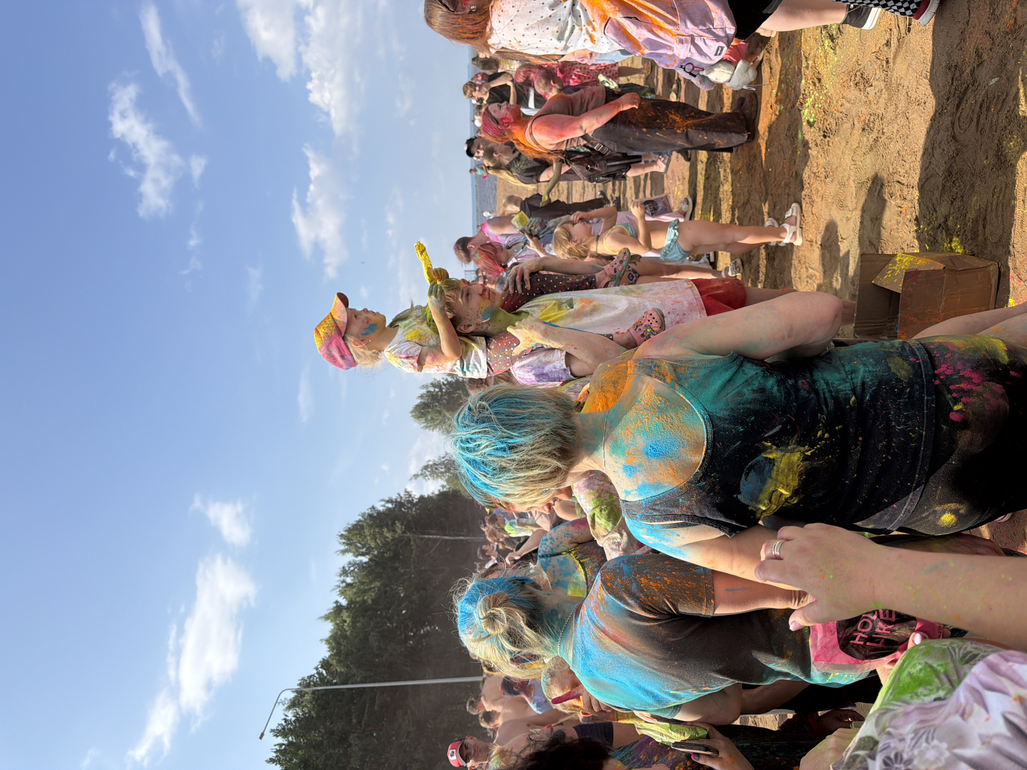 In Chaikovsky, Russia, a lively outdoor gathering is taking place under a clear blue sky, with scattered white clouds. The scene is dominated by people covered in brightly colored powders, indicative of a Holi festival celebration.
In the foreground, on the left, a person is seen from the chest down, their arm reaching out, holding a pink bag with black lettering that appears to say "HOLI LIKE". They are wearing a colorful, patterned garment. Adjacent to them, a person in a dark tank top, also covered in blue, yellow, and orange powder, is facing away from the camera. To their left, another individual with blonde hair styled in a bun, also heavily dusted with blue and orange powder, is partially visible.
Further back, a young girl wearing a yellow and pink baseball cap is perched on the shoulders of a young man. She is holding a yellow object, possibly a water gun or a pouch of colored powder. The young man, wearing a white t-shirt with red shorts, is also covered in colored powder, particularly on his face and neck.
The crowd consists of various individuals, including adults and children, many of whom have colored powder on their clothing and skin. Some people are holding bags or containers of powder, suggesting active participation in the festival. The ground is sandy or dirt, also littered with colored powders. In the background, a line of green trees separates the gathering from a body of water, likely the Kama River. A street light pole is visible on the far left, indicating the edge of a populated area. The overall atmosphere appears festive and energetic.