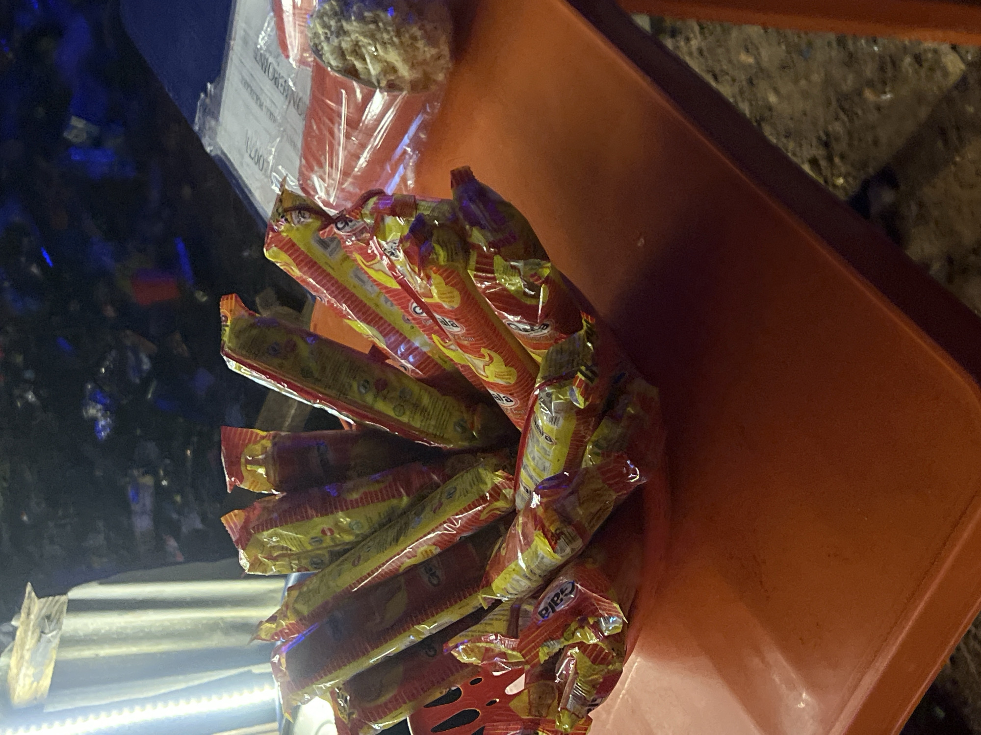 The image shows a close-up of a red bowl overflowing with individually wrapped snacks. The snacks are cylindrical and appear to be packaged in shiny red and yellow wrappers with some text visible, possibly a brand name. They are arranged in a haphazard manner, filling the bowl. The bowl itself has a decorative, possibly perforated edge, visible on the left side. The bowl is placed on an orange surface, which appears to be a table. In the background, there is a dark, blurred environment with some lights and possibly some posters or signs. To the right of the bowl, a white paper with text is visible on the orange table, and next to it, a round object, possibly a food item wrapped in plastic, is present. The lighting in the scene suggests it might be nighttime or an indoor setting with artificial lights. No people or specific landmarks of Durumi, Nigeria are visible in the image.