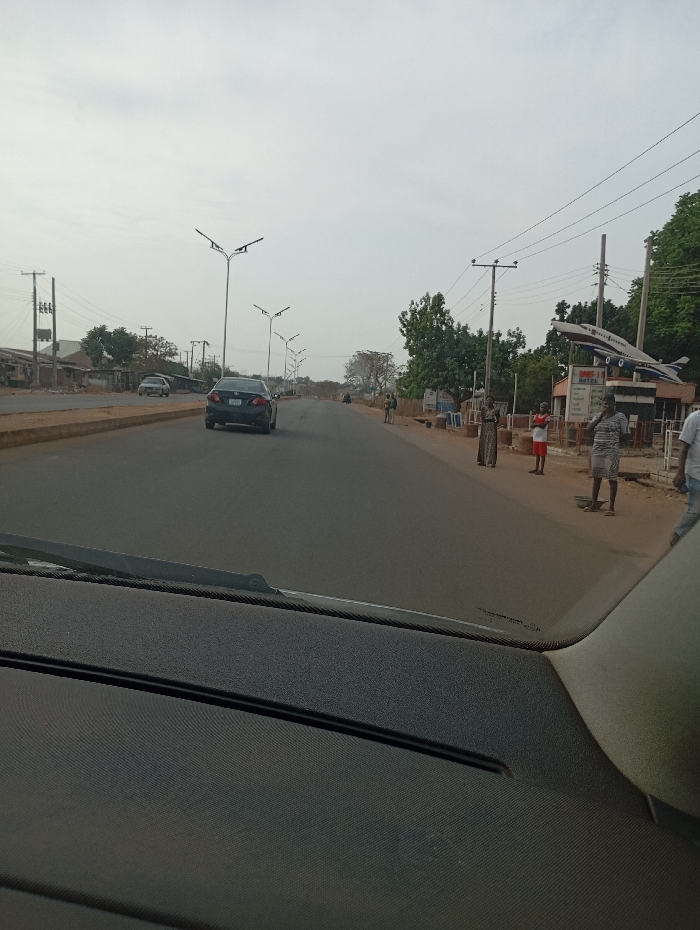 A long road stretches out under a slightly overcast sky, lined with tall streetlights. A black car drives along the road. A group of people stand on the side of the road. There is a building to the right with an airplane on top. The foreground is the dashboard of a car, from which the photo appears to have been taken. Trees are on both sides of the road.