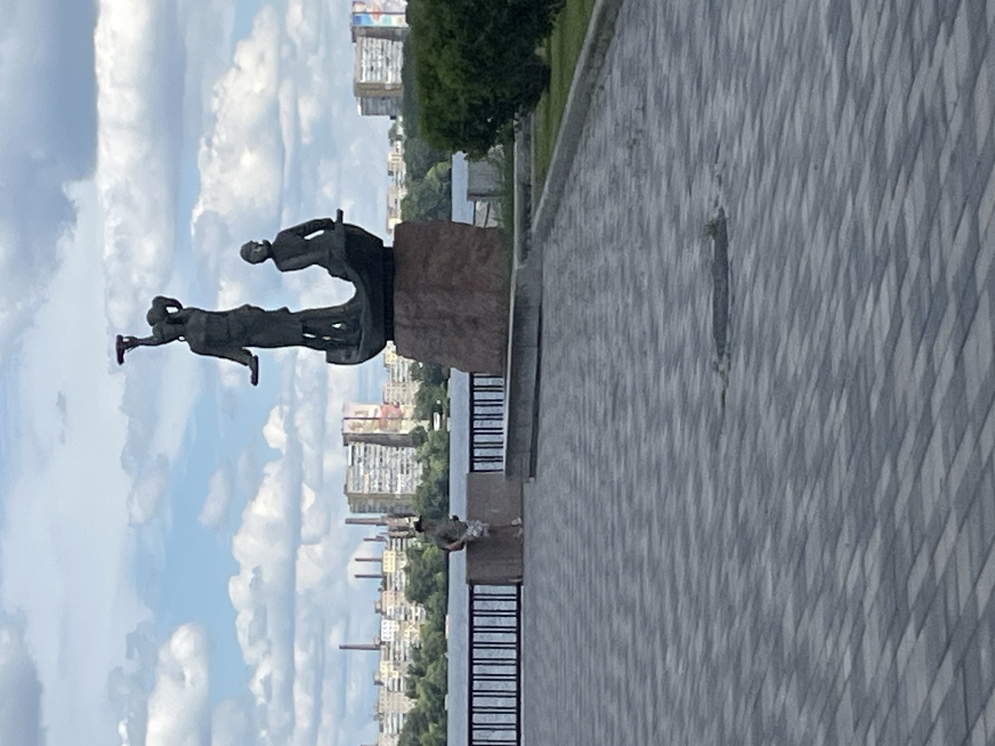 This media file is a photograph taken outdoors in Dnipro, Ukraine. The scene depicts a monument composed of two dark, abstract human figures in mid-action, seemingly connected or interacting, mounted on a reddish-brown stone pedestal. One figure appears to be reaching upwards with an outstretched arm, while the other is leaning forward.
The monument is situated on a paved walkway, characterized by a pattern of light and dark gray geometric tiles. This walkway runs alongside a body of water, likely the Dnipro River, which is visible in the background. Beyond the water, a cityscape of apartment buildings and some industrial structures, possibly chimneys, is discernible. Green trees line the riverbank, creating a contrast with the urban architecture.
The sky is filled with large, fluffy white clouds against a light blue backdrop, suggesting a bright, possibly partly cloudy day. The orientation of the photograph is vertical, with the monument appearing to the left and the walkway and cityscape extending towards the right and into the distance.
In the middle ground of the image, closer to the water's edge, there is another, smaller monument or sculpture, this one depicting a lone human figure in a crouched or kneeling posture.
The overall impression is one of a public space, possibly a park or embankment, dedicated to remembrance or artistic expression, set against the backdrop of a Ukrainian city. The time of day appears to be daytime, with ample natural light. There is no visible text in the image.