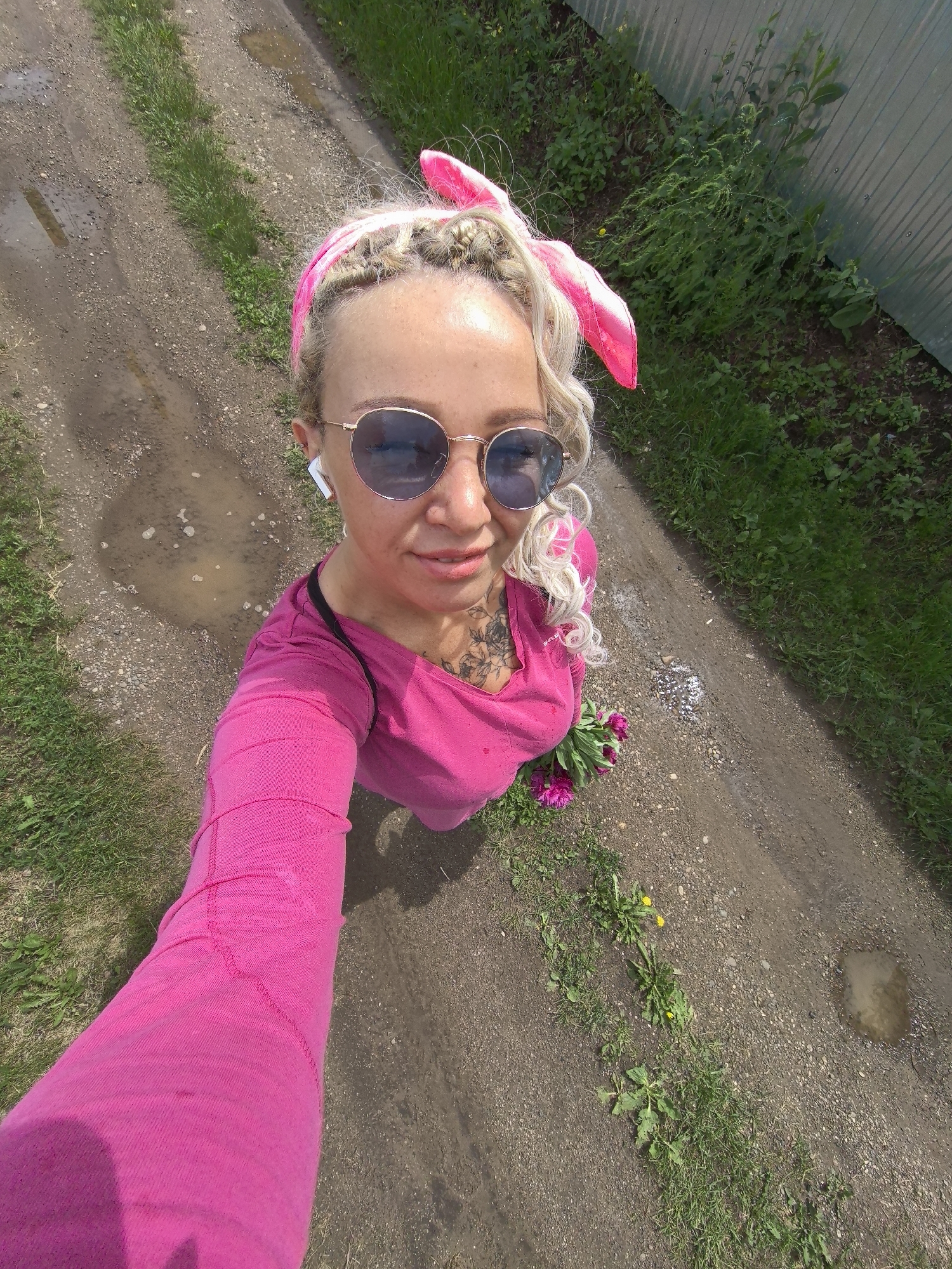 In this overhead, selfie-style shot taken outdoors in Noviy, Russia, a woman with blonde braided hair adorned with a pink bunny ear headband is captured from the chest up. She is wearing round, blue-tinted sunglasses, white wireless earbuds, and a vibrant pink long-sleeved shirt. A black strap is visible over her left shoulder. She has a tattoo on her chest that extends slightly onto her shoulder. A small bouquet of pink flowers rests near her hip.
The scene is a dirt path bordered by green grass and a corrugated metal fence. The path shows signs of recent rain, with several puddles of muddy water visible. Dandelions are scattered amongst the grass. The lighting suggests it is daytime, with bright sunshine casting shadows. The overall mood appears casual and cheerful, with the woman's slight smile and the playful headband. No visible text is present in the image.