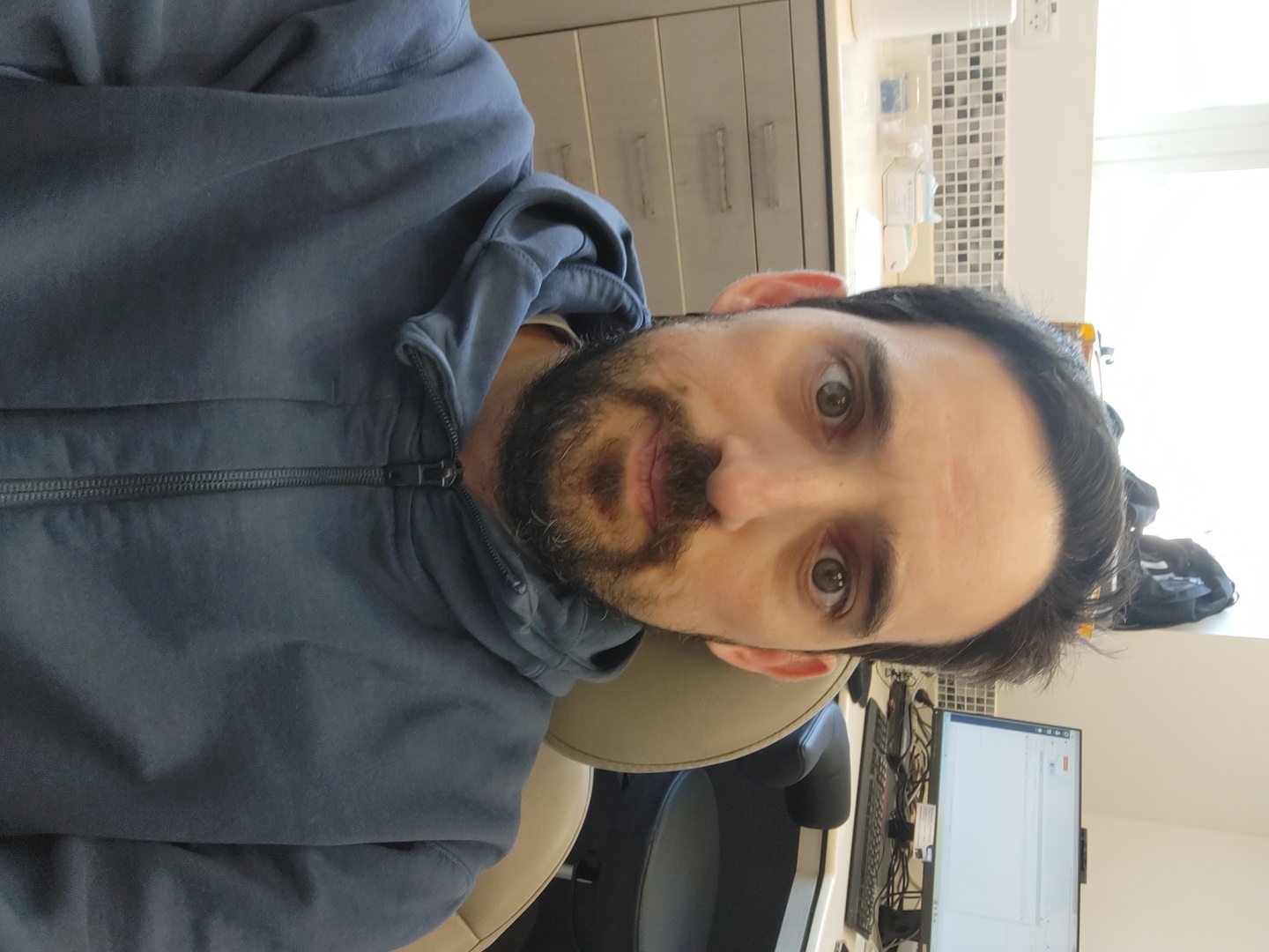 A man with dark hair and a trimmed beard, wearing a blue hooded sweatshirt, is sitting in an office. A computer monitor and keyboard are visible on a desk in front of him. Behind him is a file cabinet and a tiled wall. The location is likely Tel Aviv, Israel, based on the visible European power outlet.