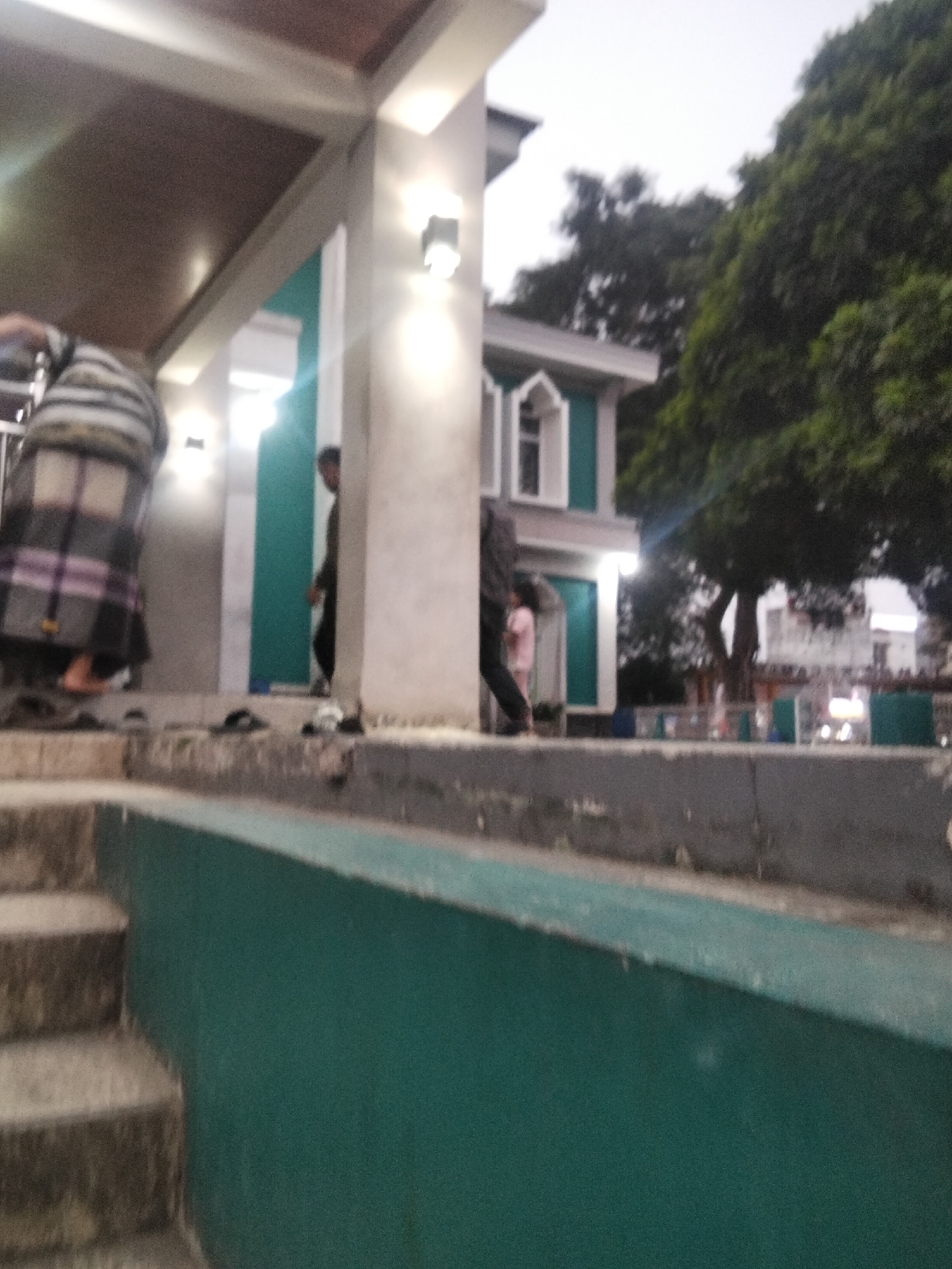 The image depicts an outdoor scene, possibly near a religious building, with a hint of what appears to be the city of Cisaat, Indonesia. In the foreground, a set of concrete steps leads up to a structure with a teal-colored wall. To the left, a person is visible from the waist down, wearing a plaid sarong. Further into the scene, there is a building with green accents and white trim, and a large tree dominates the right side of the frame. In the background, the sky is overcast. The overall atmosphere is subdued, with artificial lighting visible on the building's exterior.