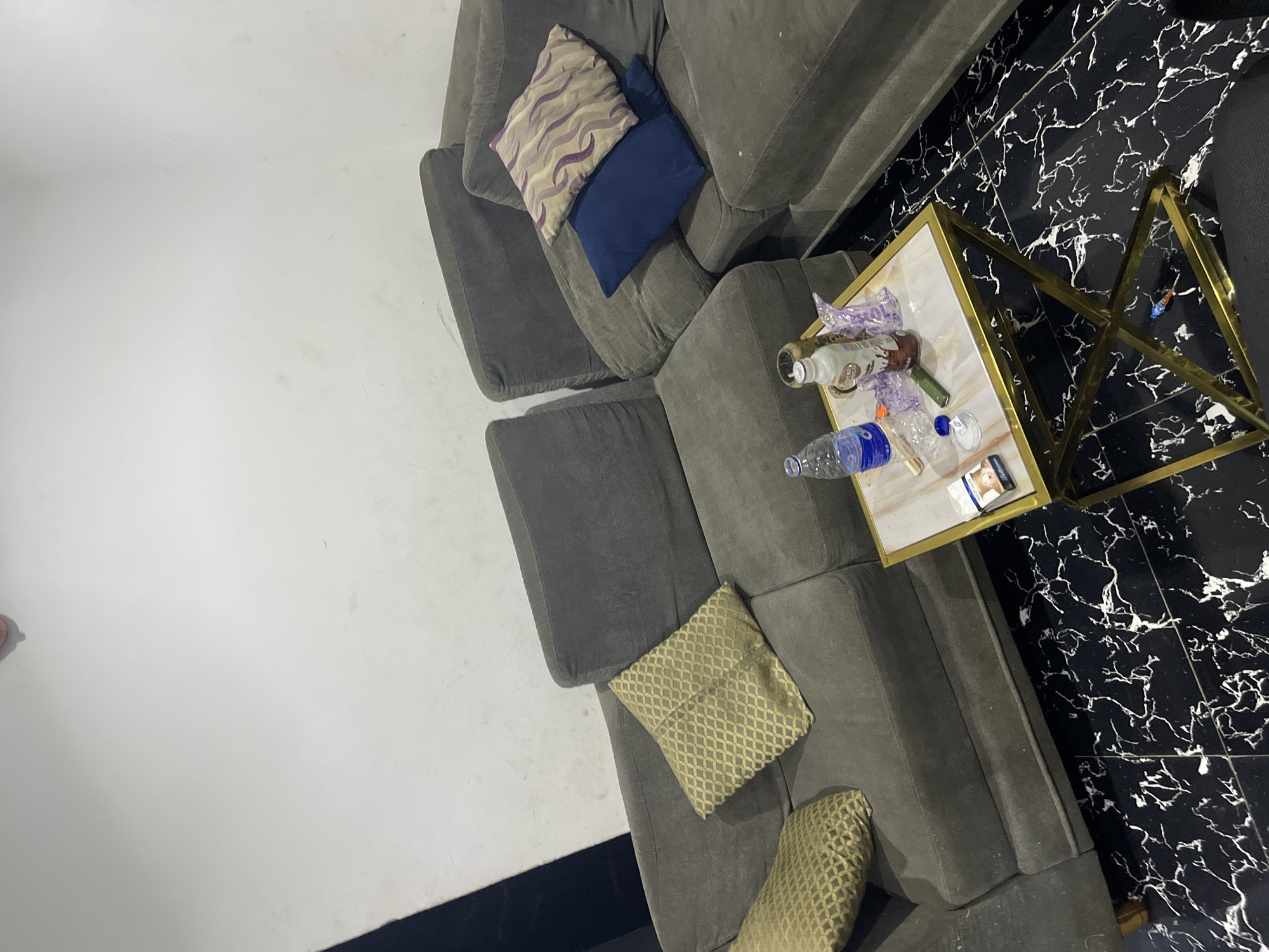 A high-angle shot shows a section of a grey, sectional sofa with several pillows, including two geometric print throw pillows, one purple and tan patterned pillow, and one navy blue pillow. Adjacent to the sofa is a white-topped coffee table with a gold frame, positioned on a black and white marble-patterned floor. Several items are on the table, including two plastic water bottles, an empty glass bottle, various wrappers, a cigarette pack, and a small white container. The photo is taken indoors, with a white wall visible in the background. No people are visible in the image. There is no definitive information in the image to confirm it is in Abuja, Nigeria.