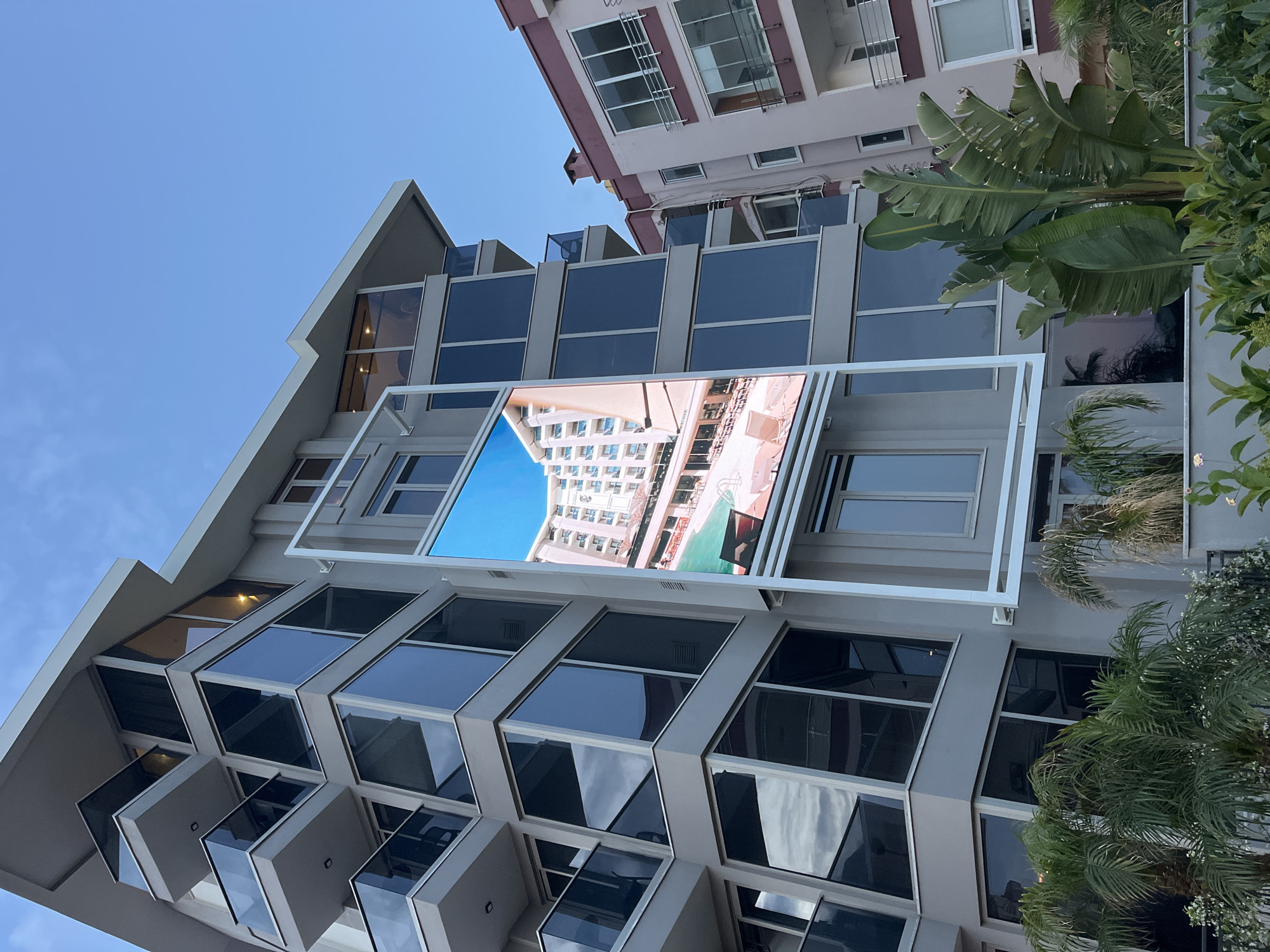 A low-angle, upward-tilted shot captures the facade of a building in Alanya, Turkey, featuring distinctive contemporary architecture. The building's exterior is primarily gray with large, rectangular windows that reflect the sky. The windows are arranged in a staggered pattern, creating a dynamic visual effect. A rectangular white frame is mounted on the building's facade, displaying an image that reflects another building with a swimming pool and blue sky.
To the right of the main building, part of another building is visible. It has a light pink facade and white-framed windows. Palm trees and other greenery are interspersed around the buildings. The sky is a clear, light blue.