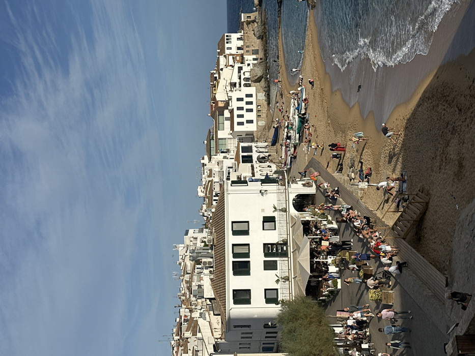 This image captures a panoramic view of a coastal village in Calella De Palafrugell, Spain, under clear, sunlit conditions. The scene is dominated by a cluster of white-washed, multi-story buildings with dark tiled roofs, ascending a gentle hillside from the shoreline.
In the foreground and midground, a paved promenade transitions into a wooden boardwalk, separating the buildings from a crescent-shaped sandy beach. A prominent building with a curved corner, dark green shutters, and a lower-level veranda supporting an outdoor restaurant occupies the left-center. This restaurant features numerous tables, chairs, and large beige or white parasols, under which approximately two dozen patrons are seated and interacting.
The promenade and boardwalk are populated by numerous individuals, estimated at 30-40, casually dressed, walking, standing, and observing the scene. A wooden staircase descends from the boardwalk directly onto the beach.
The sandy beach, extending along the right side of the image, is occupied by approximately 20-30 individuals engaged in leisure activities. Several small boats, predominantly white, blue, and green, are pulled onto the sand. People are seen sunbathing on towels, walking along the water's edge, and one individual appears to be bending over in the sand. The water is a clear blue-green, with small waves breaking gently on the shore. Rock formations are visible at the far right edge, marking the end of the beach.
The sky is a pale blue with scattered, wispy cirrus clouds. The bright sun, likely late afternoon or early morning, casts distinct shadows across the promenade, beach, and parts of the buildings. The overall impression is one of a vibrant, active Mediterranean coastal town.