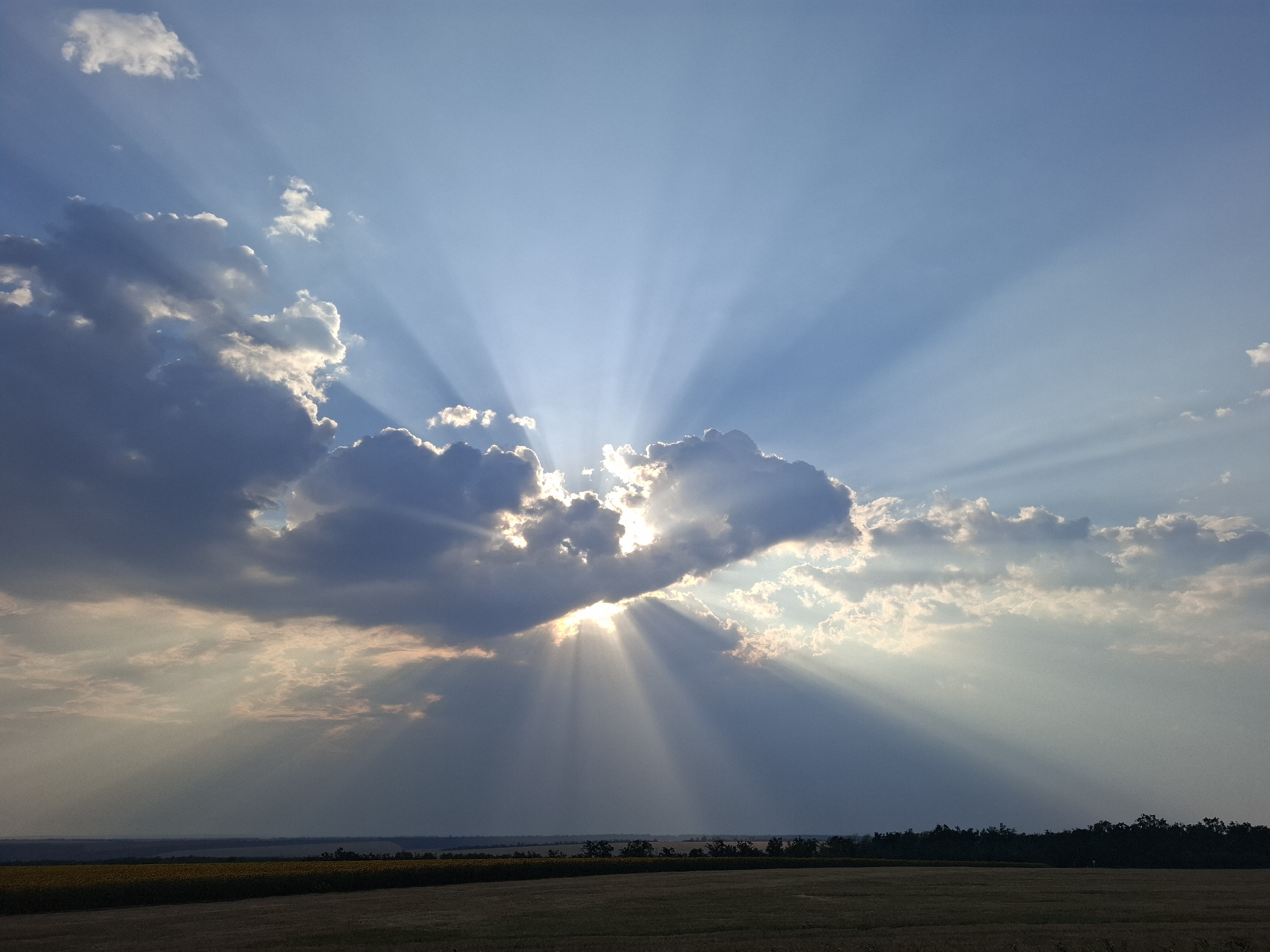 The image shows a dramatic sky with crepuscular rays emanating from behind a cloud formation. The sun is partially obscured by a dark, textured cloud in the center of the frame, creating a radiant effect as its light beams pierce through the atmosphere. The sky is a vibrant blue, transitioning to a lighter shade near the horizon. Below the sky, a landscape is visible, featuring a dark silhouette of trees and a field. The foreground consists of a field with hints of yellow and brown, suggesting agricultural land. The overall scene captures a moment of intense natural light and atmospheric activity, likely during sunrise or sunset, over the rural area of Topaly, Ukraine. There are no people or specific actions depicted in the image.