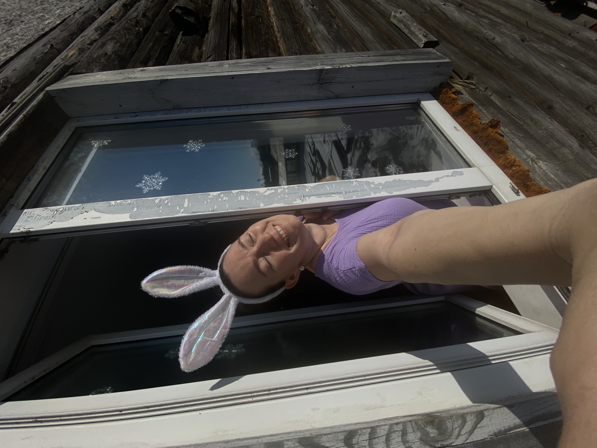A woman wearing white bunny ears looks out of a window. The window is in a wooden structure. Snowflake decorations are on the window. The location is Ольховка, Russia.