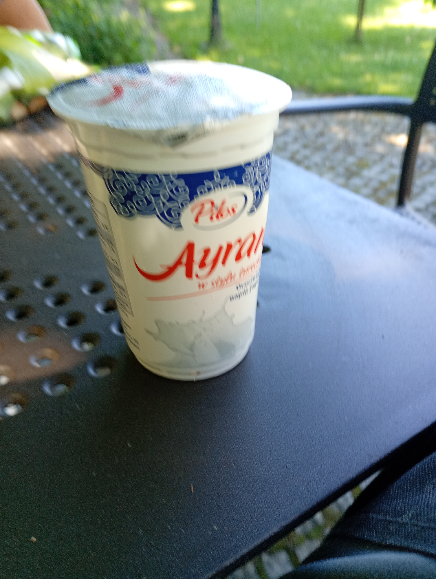 A close-up, slightly blurry shot captures a single white yogurt cup with a foil lid. The cup features a blue and white decorative border around the top, and bold red lettering that spells out "Ayran". A small logo with the word "Pilos" is visible above "Ayran". The cup sits on a dark, textured surface that appears to be an outdoor table, suggested by the dappled sunlight and the out-of-focus greenery in the background. A hint of blue denim fabric is visible in the lower right corner, possibly indicating the presence of a person. The scene suggests a casual outdoor setting, perhaps a park or patio, on a bright day. The focus is on the yogurt cup, implying it is the primary subject of interest. The overall impression is a simple, everyday moment captured in an outdoor environment.