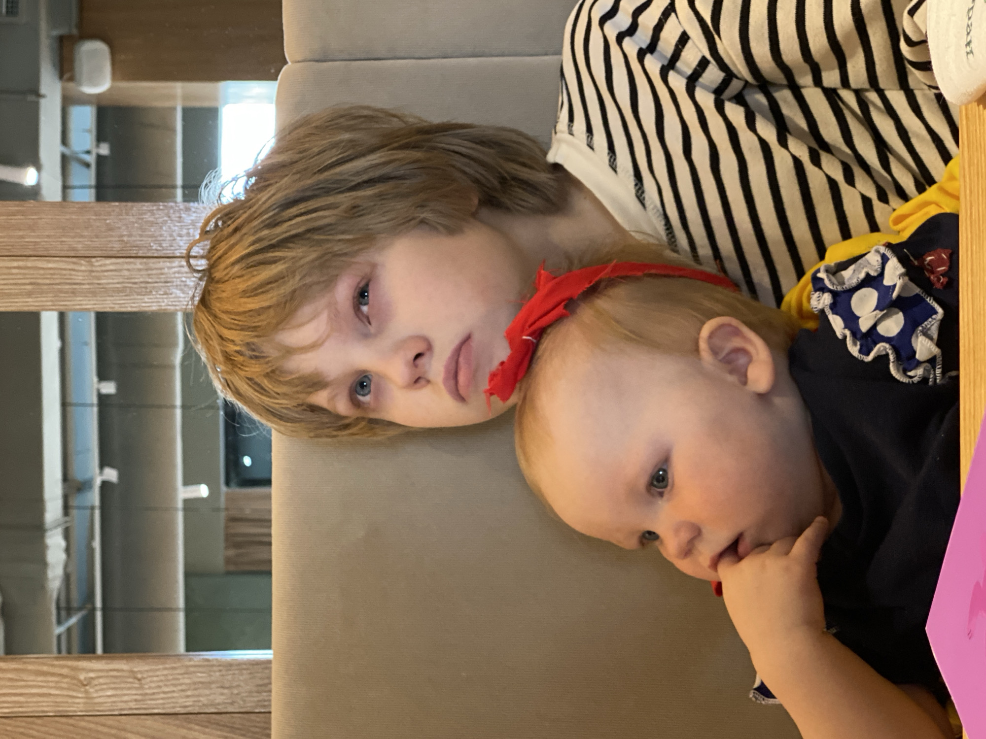 Two young children are lying down together on a beige upholstered surface, likely a sofa or bench. The older child, with light brown hair and blue eyes, is lying on their back with a somewhat somber expression. Resting near the older child is a baby wearing a red ribbon or bow, who has fair skin, light hair, and is looking downwards while sucking their thumb. Both children are wearing casual clothing. In the background, there's a wall with decorative vertical wooden slats and a glimpse of a building exterior through the window, indicating an interior location. Based on the information provided, I am unable to confirm if this location is in Yekaterinburg, Russia.