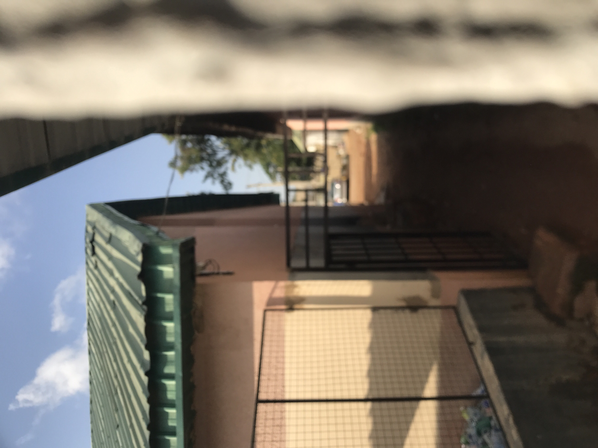 The image, captured in New Stadium, Nigeria, depicts a narrow outdoor passage or alleyway under a partly cloudy blue sky. On the left, a building with a green corrugated sheet metal roof and a light peach-colored wall is visible. In front of the wall, a black metal mesh fence rests on a low concrete base, behind which scattered debris, possibly plastic bottles and bags, is present. A black metal gate further along the reddish-brown unpaved pathway creates a narrower access point. In the distant background, a light-colored vehicle is faintly discernible near green foliage. The entire right foreground is dominated by a large, blurred, light-toned vertical structure, likely a pillar or wall, indicating extreme proximity to the camera. The scene is uniformly soft-focused throughout. No human figures or specific actions are discernible.