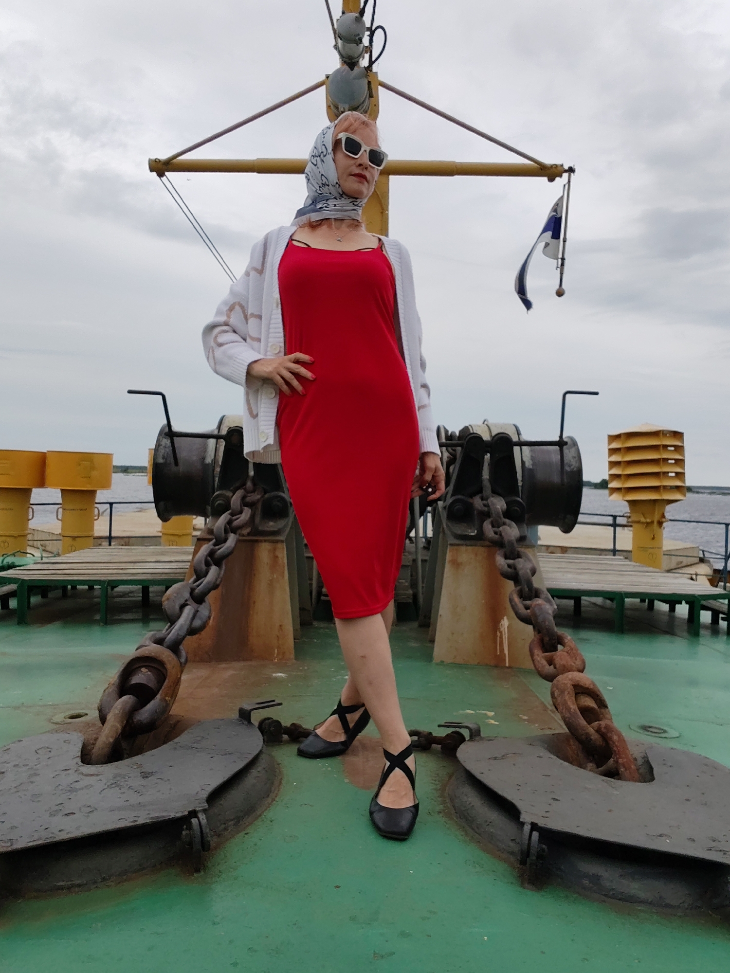 A woman stands on the deck of a ferry with her left hand on her hip and her right hand resting near her waist. She is wearing a red knee-length dress, a white cardigan with a heart pattern, a patterned headscarf, white sunglasses, and black flats with ankle straps. The ferry deck is green and appears wet. In the foreground, a large anchor chain is visible. In the background, there are two large anchor winches and yellow structures on the sides of the ferry. A flag on a pole is also visible. The sky is overcast. The location is the city of Novokemskii, Russia.