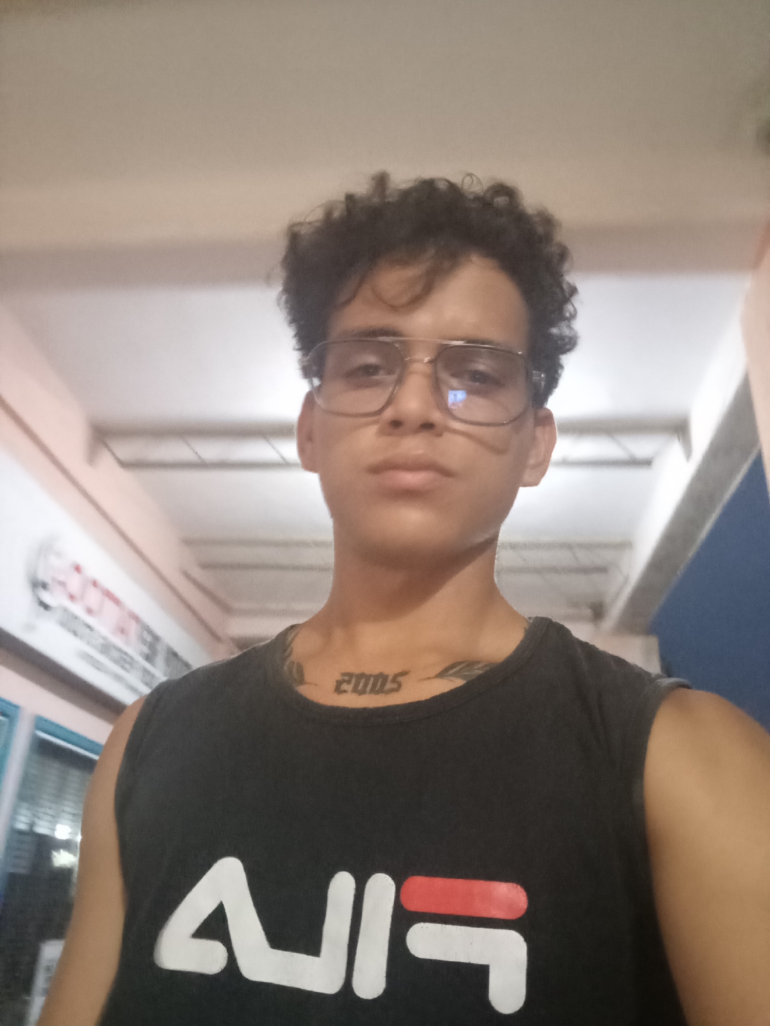 A young man with curly dark hair and glasses is captured in a selfie. He is wearing a black sleeveless t-shirt with a white and red logo. A tattoo is visible on his neck, with the number "2005" prominently displayed. He appears to be indoors, with a ceiling and some background structures visible. The setting is likely a commercial establishment, possibly a gym or sports store, given the signage in the background which includes text that might be "FOOTATTACK". The lighting is artificial and somewhat dim. The overall impression is that of a casual, personal snapshot taken in an urban environment.