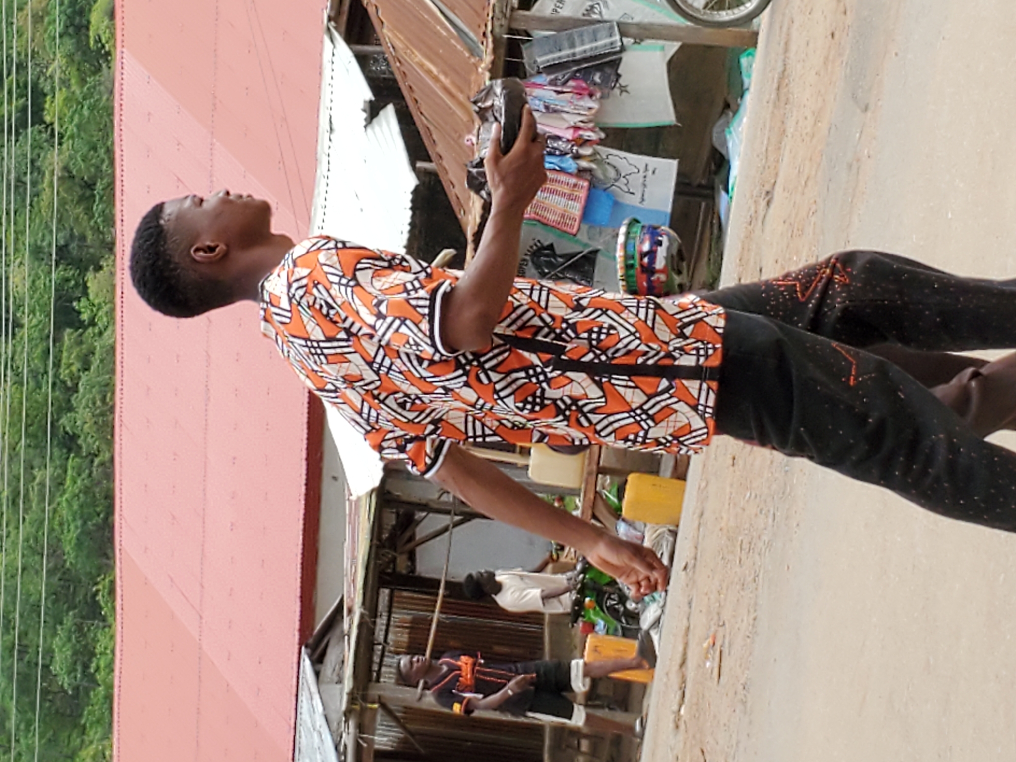 A street scene in Mararaba, Nigeria, shows a young man in an orange and black patterned shirt and dark pants standing on a sidewalk. He is holding a dark, rectangular object in his right hand and looking up. Behind him is a building with a reddish-brown roof. Several other individuals are visible in the background, along with a variety of objects including a bicycle wheel, some containers, and some merchandise. The area suggests a roadside business or market environment. Trees and utility lines are visible in the background. The ground is light-colored, potentially concrete or dirt.