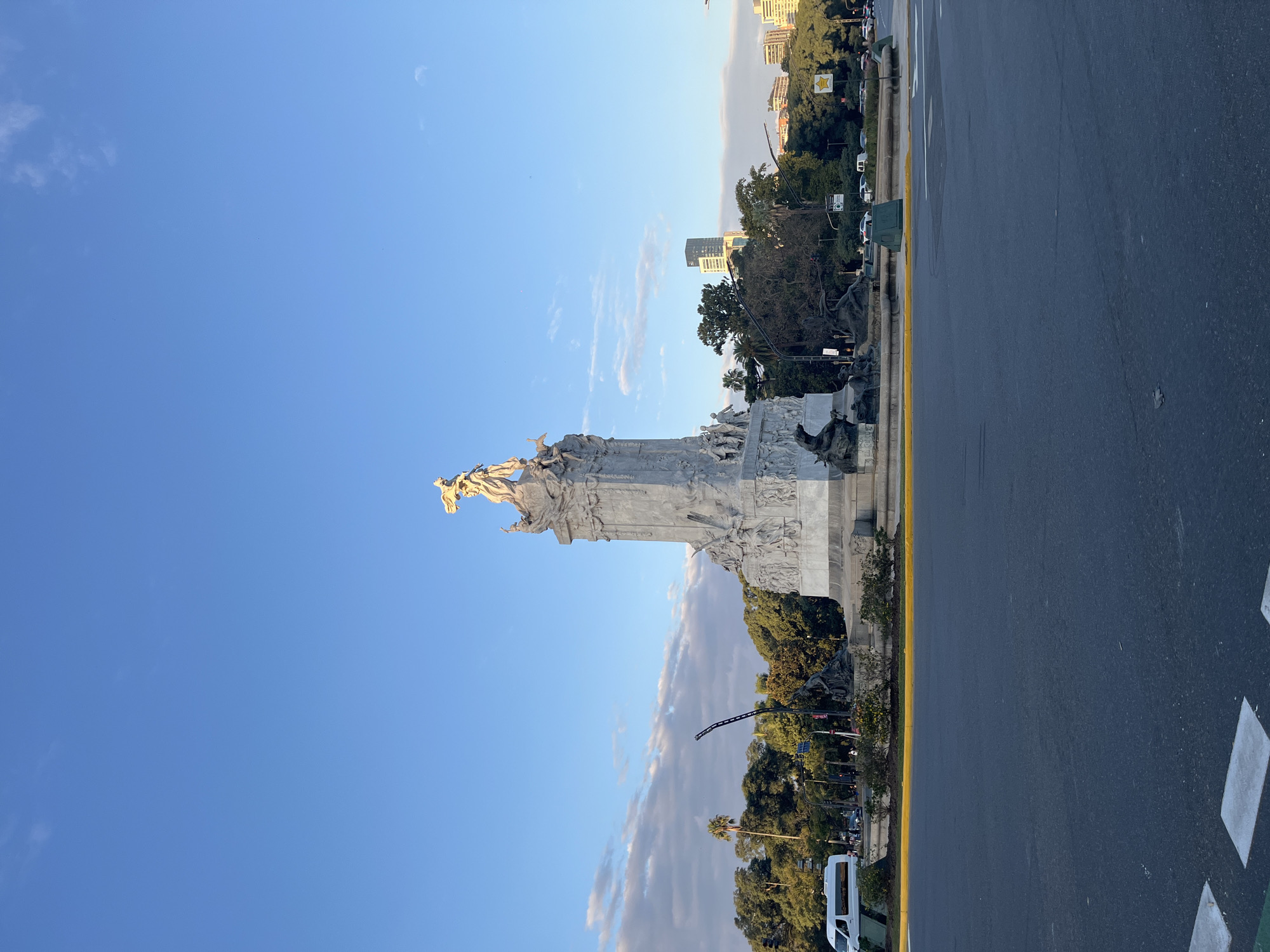 The image shows the Monument to the Carta Magna and Four Regions of Argentina, commonly known as the "Monument of the Spanish", in Buenos Aires, Argentina. The monument is made of white marble and features sculptures of various figures and allegories. The image is taken from a low angle, emphasizing the monument's height against a bright blue sky with some clouds. Next to the monument is a street with a yellow curb. In the background, buildings and trees can be seen.