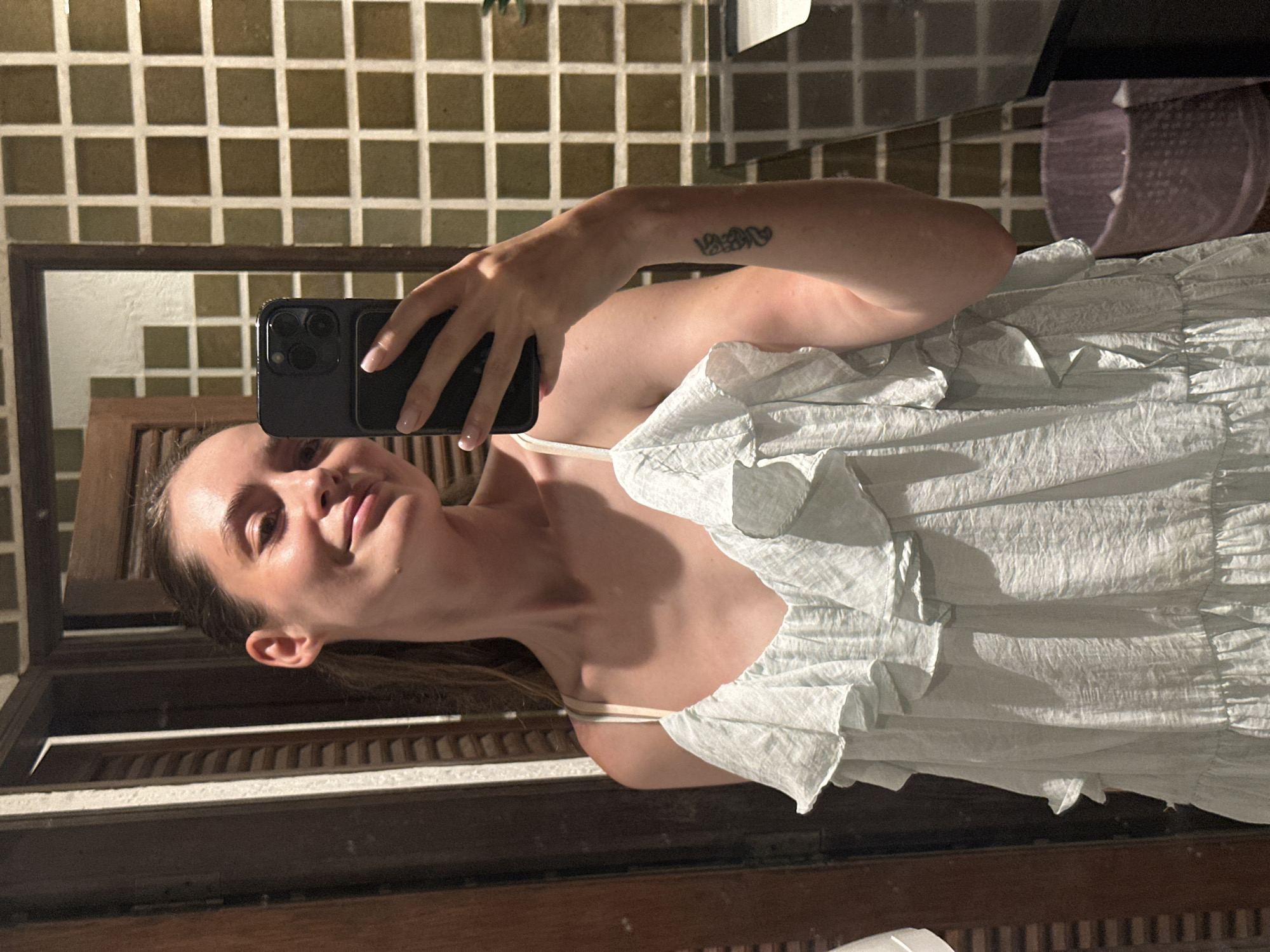 A woman is taking a selfie in a bathroom. The bathroom has tan tile walls and a brown-framed mirror. The woman is wearing a light blue ruffled dress with spaghetti straps. She is holding a black iPhone in her left hand. The woman has a tattoo on her left forearm. There is a brown wooden surface near the bottom of the photo. A gray item is in the upper right of the photo. This is likely in Phuket City Municipality, Thailand.
