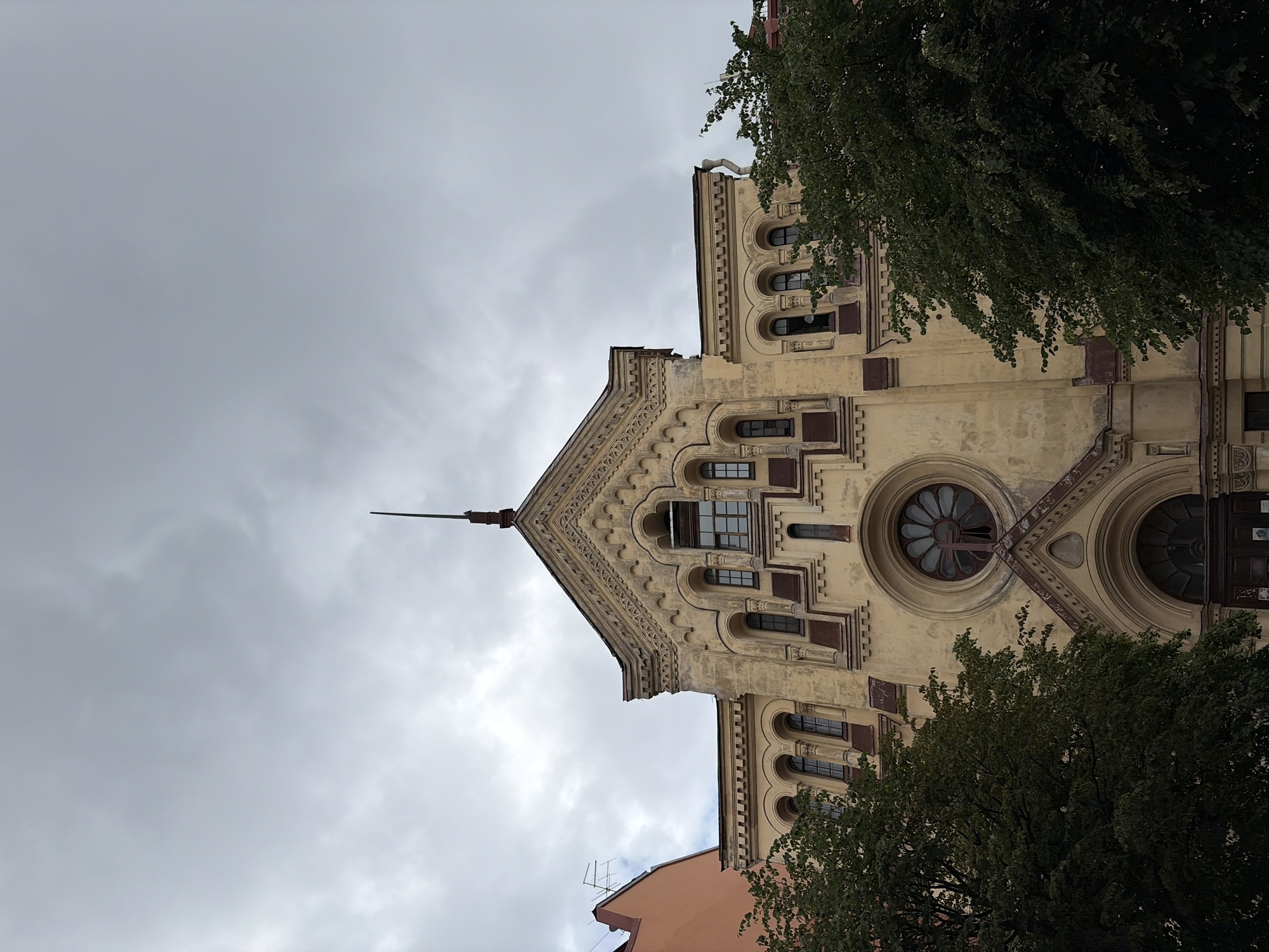 The image shows the facade of the Great Choral Synagogue in Saint Petersburg, Russia, under a cloudy sky. The yellow building features ornate architectural details, including arched windows, decorative carvings, and a prominent rose window with a cross inside. Trees frame the lower portion of the building, obscuring part of the entrance. The sky is overcast and gray, indicating a potentially inclement weather day. There are no people visible in the frame.