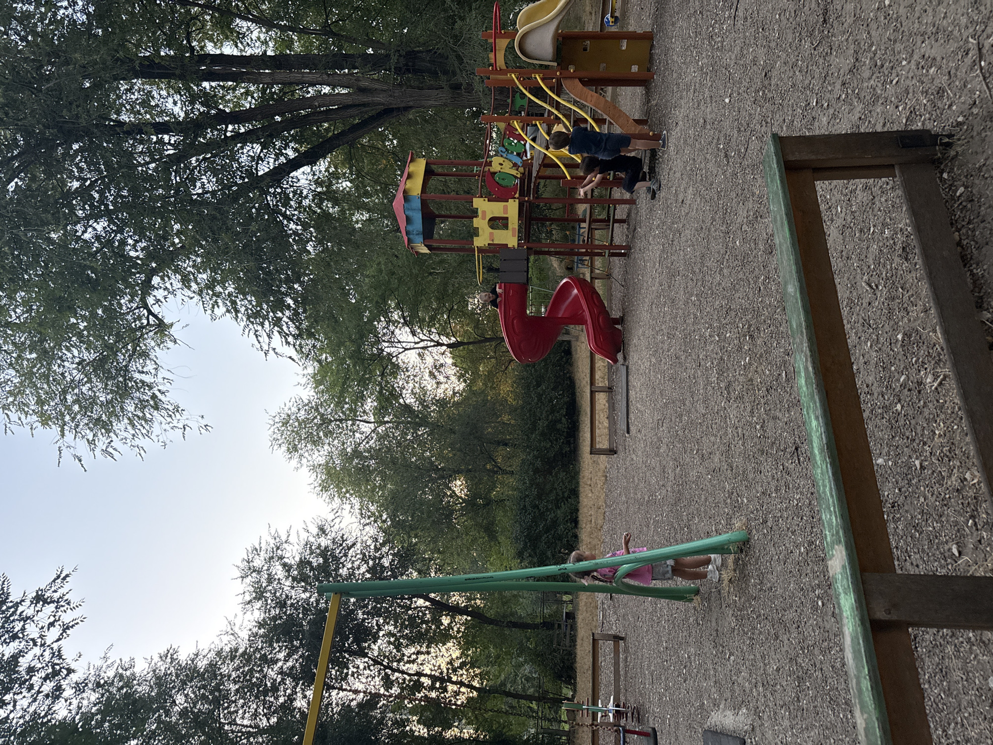 The image depicts a playground scene at dusk in Chișinău, Moldova. A young girl wearing a pink shirt and grey shorts stands near a green swing set in the foreground. In the background, a colorful playground structure with slides and climbing elements is visible. Several children are interacting with the playground equipment. The ground is covered with gravel, and mature trees surround the area, suggesting a park or recreational space. The sky is pale, indicating the end of the day.