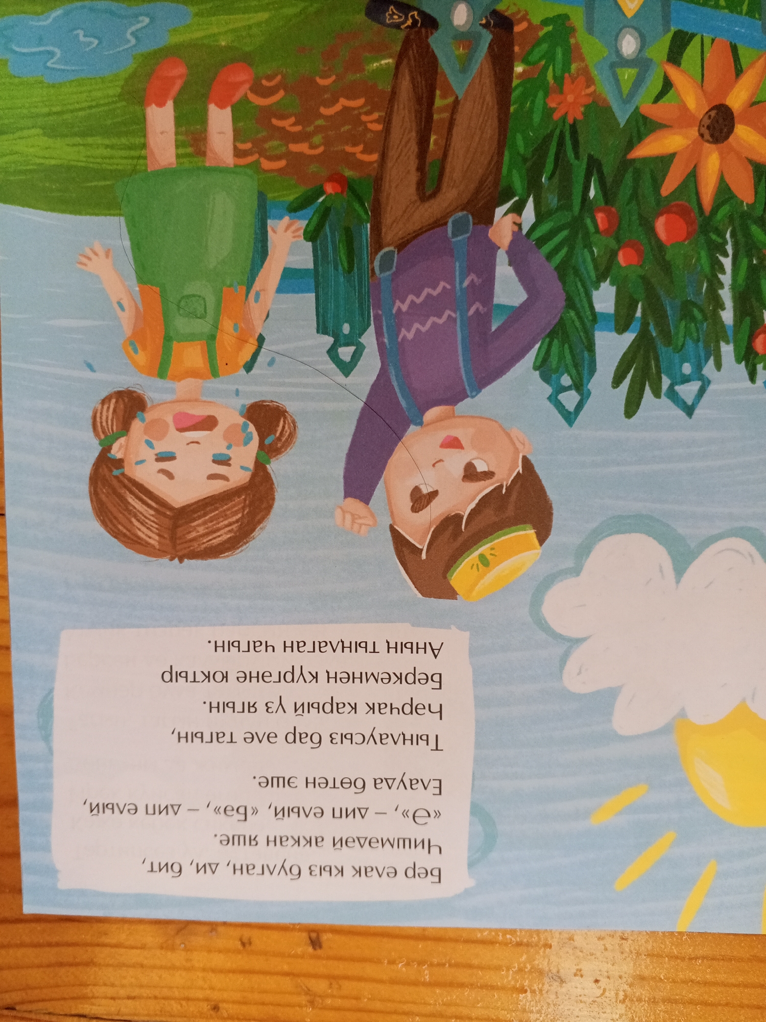 The image depicts a colorful illustration, possibly from a children's book, with several children in an inverted orientation, suggesting they are upside down. On the left, a child in a green backpack and orange overalls with red shoes is shown with their legs in the air. Next to them, a crying child with brown hair in two buns and blue markings on their face is also inverted. To the right, another inverted child wearing brown pants and a purple top with suspenders is depicted. The background features a stylized depiction of nature with green foliage, orange flowers, red fruit, and a bright yellow sun. Below the illustration, there is text written in a script that appears to be Cyrillic. The image does not contain any elements that would allow for the identification of a specific location in Chistopol, Russia.