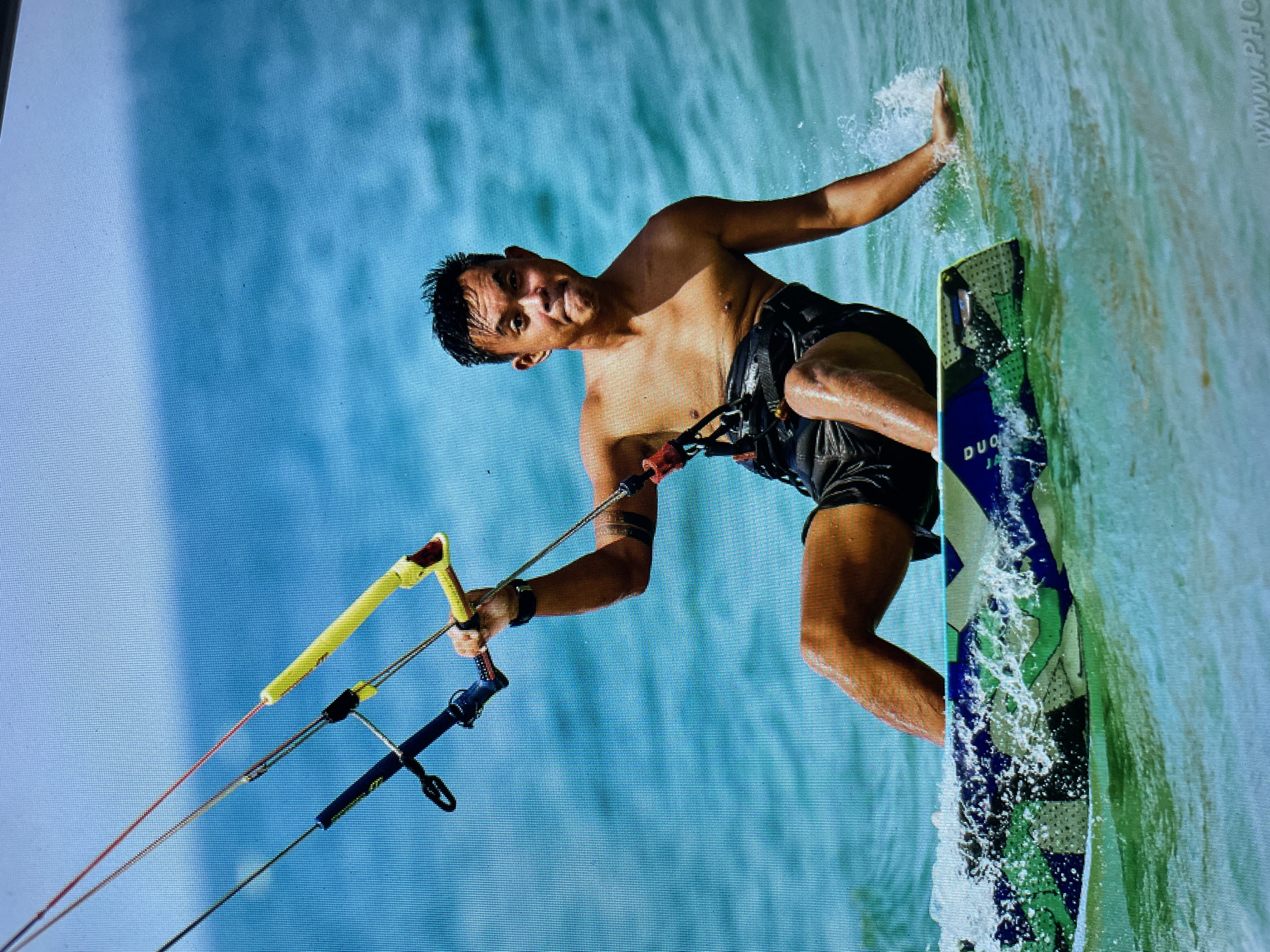 The image depicts a man kitesurfing. He is shirtless, wearing black shorts and a harness. He holds a kite bar with yellow and black elements and leans back as he rides the kiteboard through the water. The board is partially submerged and decorated with blue, green, and white designs. The background shows clear blue water. The setting is consistent with a coastal location in Bacong, Philippines.
