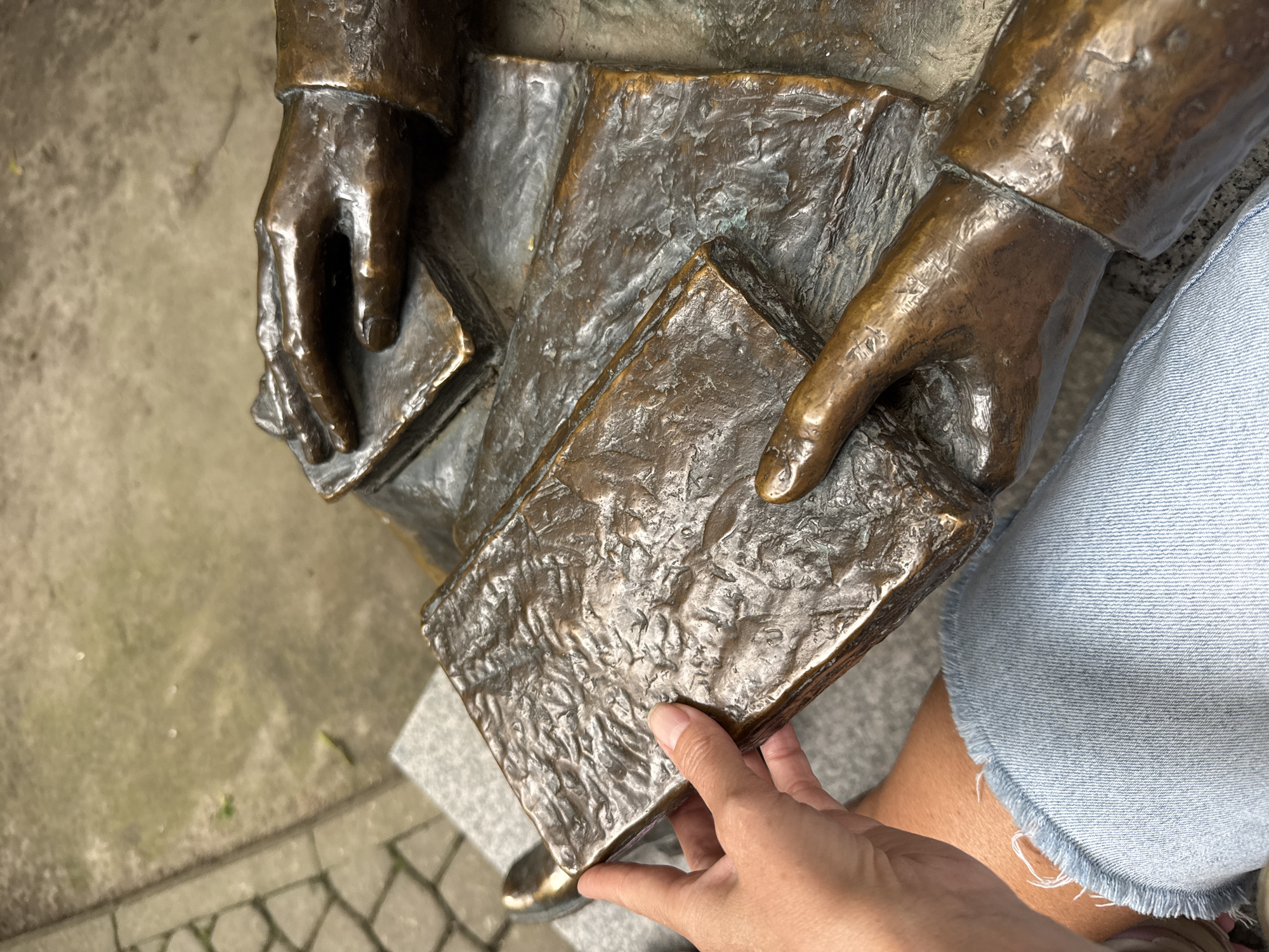 A close-up shot shows a person's hand holding a textured, bronze book. The hand, with visible fingernails, is positioned on the lower left side of the image. The book has a weathered appearance, suggesting age or a deliberate artistic choice. To the right of the hand and book, two bronze sculpted hands rest on other bronze books, one on top of the other. The sculpted hands appear to be part of a larger statue, possibly depicting a seated figure engaged in reading. The background consists of a stone-paved area and a gray granite bench. The person's legs, clad in denim shorts with frayed edges, are visible in the lower portion of the frame. The scene appears to be outdoors, possibly in a public space.