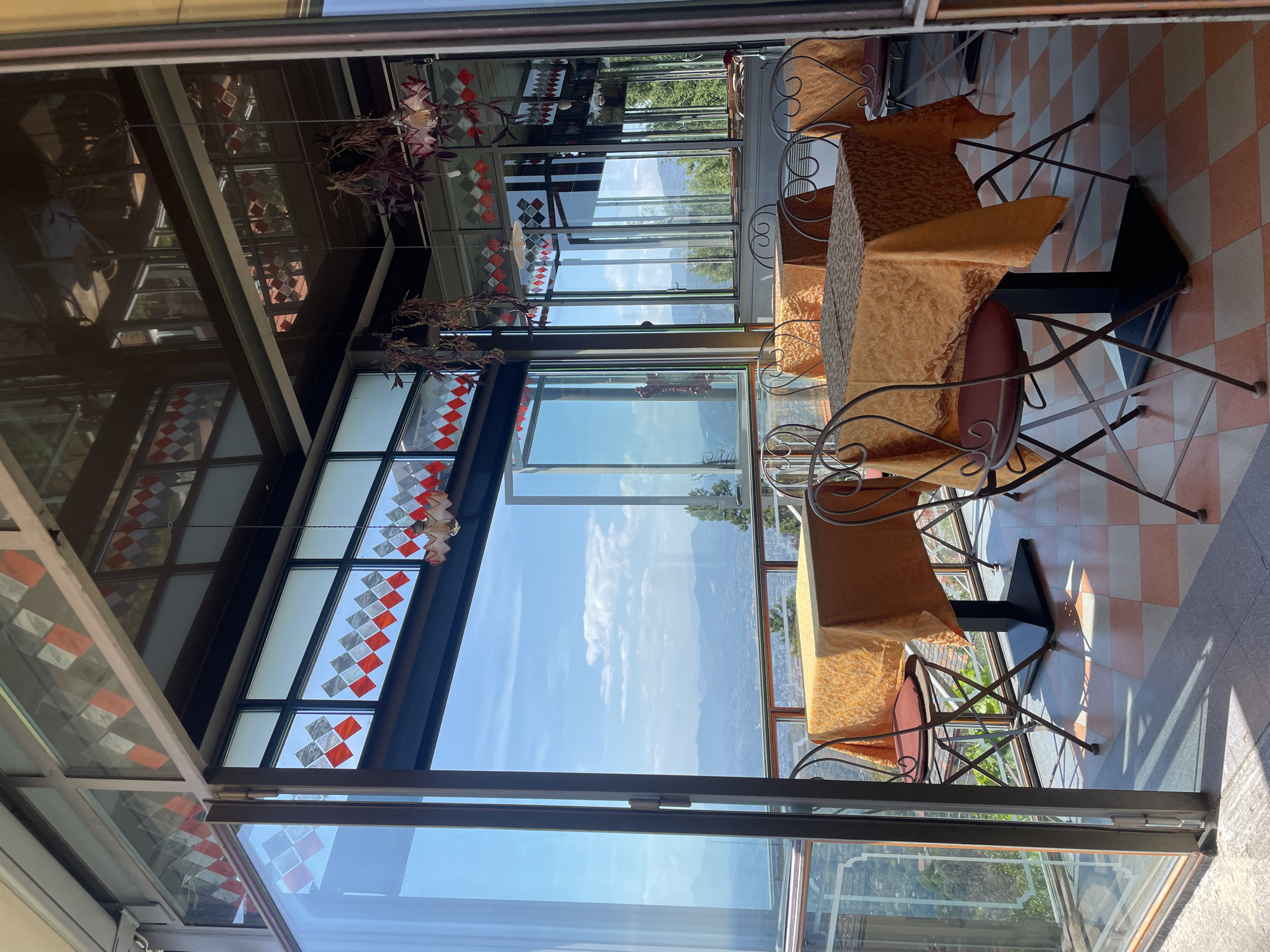 This media file shows an indoor dining area with a panoramic view of a mountainous landscape. The room features tables draped with orange tablecloths and metal chairs. The walls and ceiling are a combination of glass, metal, and decorative stained glass with diamond patterns in red and white. Plants hang from above. The large windows offer a clear view of a bright, sunny day with blue skies and scattered clouds, suggesting it is daytime. The mountains in the distance are lush and green, hinting at a natural, possibly rural or semi-rural, setting. The checkered floor inside also adds to the aesthetic of the dining space. The overall impression is one of a restaurant or cafe with a scenic overlook.