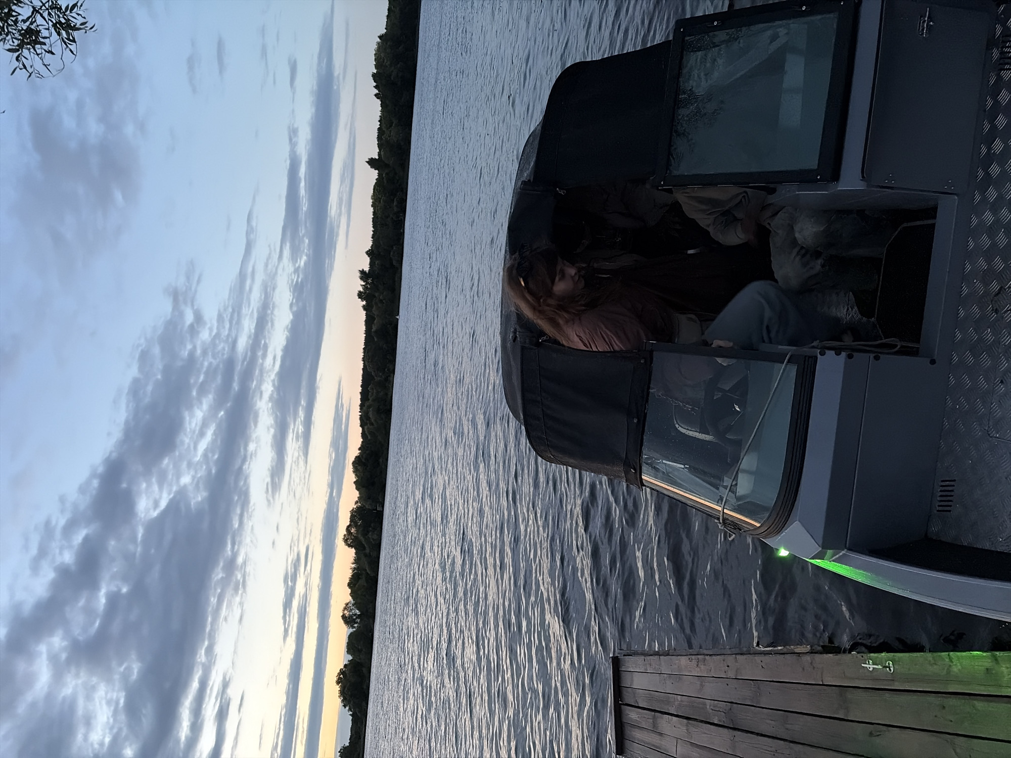 At dusk, a motorboat is docked at a wooden pier on a wide river. The sky is overcast with scattered clouds, and the water is calm with gentle ripples. The boat, which appears to be a modern aluminum vessel, is gray with a black canopy. A woman with long, auburn hair is visible inside the boat, leaning forward. Another person, mostly obscured, is seated next to her. A green light is illuminated on the front of the boat. The background shows a dense line of trees along the riverbank, with a hint of a pale orange and yellow sunset glow on the horizon. The location is the city of Novomelkovo, Russia.