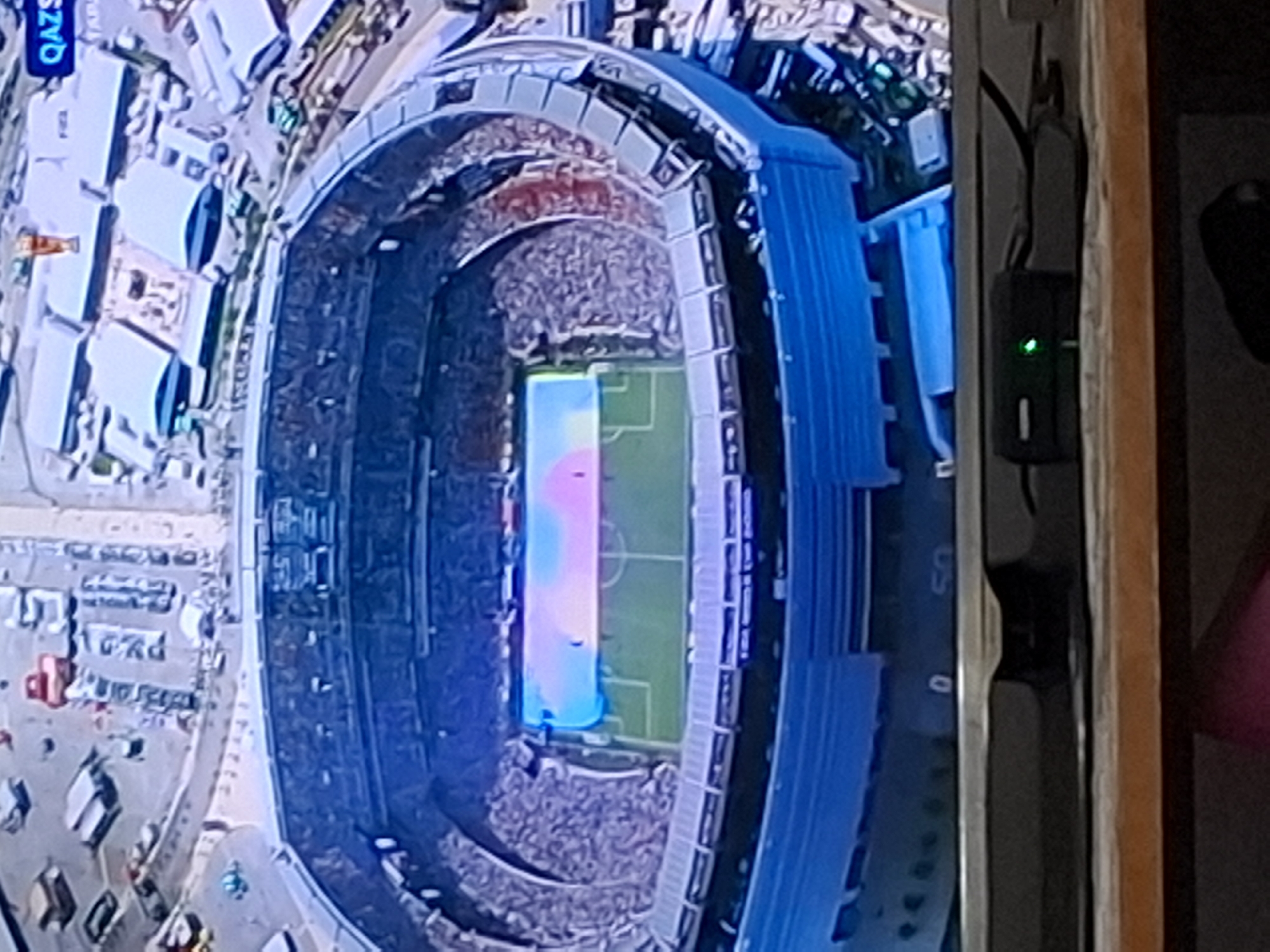 An aerial view shows a stadium in Almaty, Kazakhstan, with a colorful projection displayed on the soccer field. The stadium has blue seating and is surrounded by buildings, parking lots, and roads. The projection on the field features a blend of blue and pink hues, creating a gradient effect. In the foreground, a dark shelf holds a small black electronic device with a green indicator light. The scene appears to be captured from a high vantage point, possibly from a drone or aircraft.