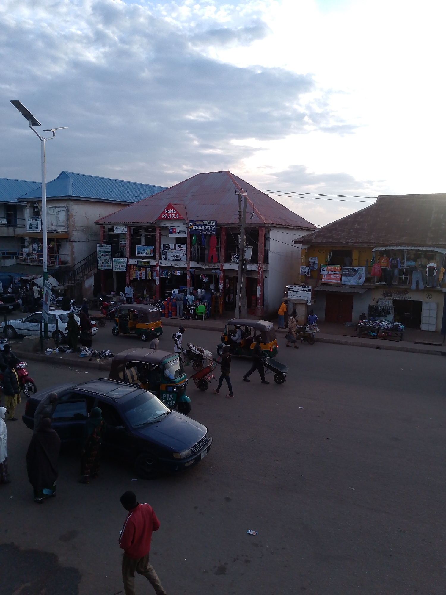 This is an aerial photograph capturing a bustling street scene in Bukuru, Nigeria, during what appears to be late afternoon or early evening, judging by the soft lighting and the presence of a street lamp. The sky is partly cloudy, with a mix of grey clouds and bright patches indicating the sun.
The scene is dominated by a street lined with several buildings, some of which appear to be shops or commercial establishments. One prominent building has a red roof and a sign that reads "NaNa PLAZA". Another building to the right has a yellow facade and displays clothing on its upper level. In front of these buildings, a busy street is filled with various modes of transportation, including several auto-rickshaws (also known as Keke Napep) and a dark-colored car. People are seen walking and interacting on the street and sidewalks.
Activities include people entering and exiting the car, individuals pushing a wheelbarrow, and pedestrians walking. The overall atmosphere suggests a lively marketplace or commercial hub. Visible text includes "NaNa PLAZA", "BJC", "NEMOMORE Collection", and other signs on the shop fronts that are difficult to fully decipher from this angle. The setting appears to be a typical urban environment in Nigeria, characterized by its vibrant street life and mix of commercial activity.