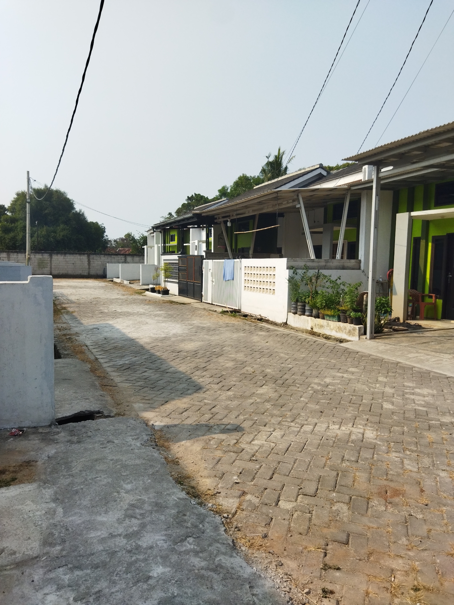 The image shows a street in the city of Suka Sari, Indonesia. The street is paved with interlocking brick pavers and concrete. On the right side of the street are several modern houses with green and white facades and corrugated metal roofs. There are also white fences, potted plants, and a red chair visible. On the left side of the street are concrete structures and a wall. The sky is clear and bright. There are no people or animals in the image.