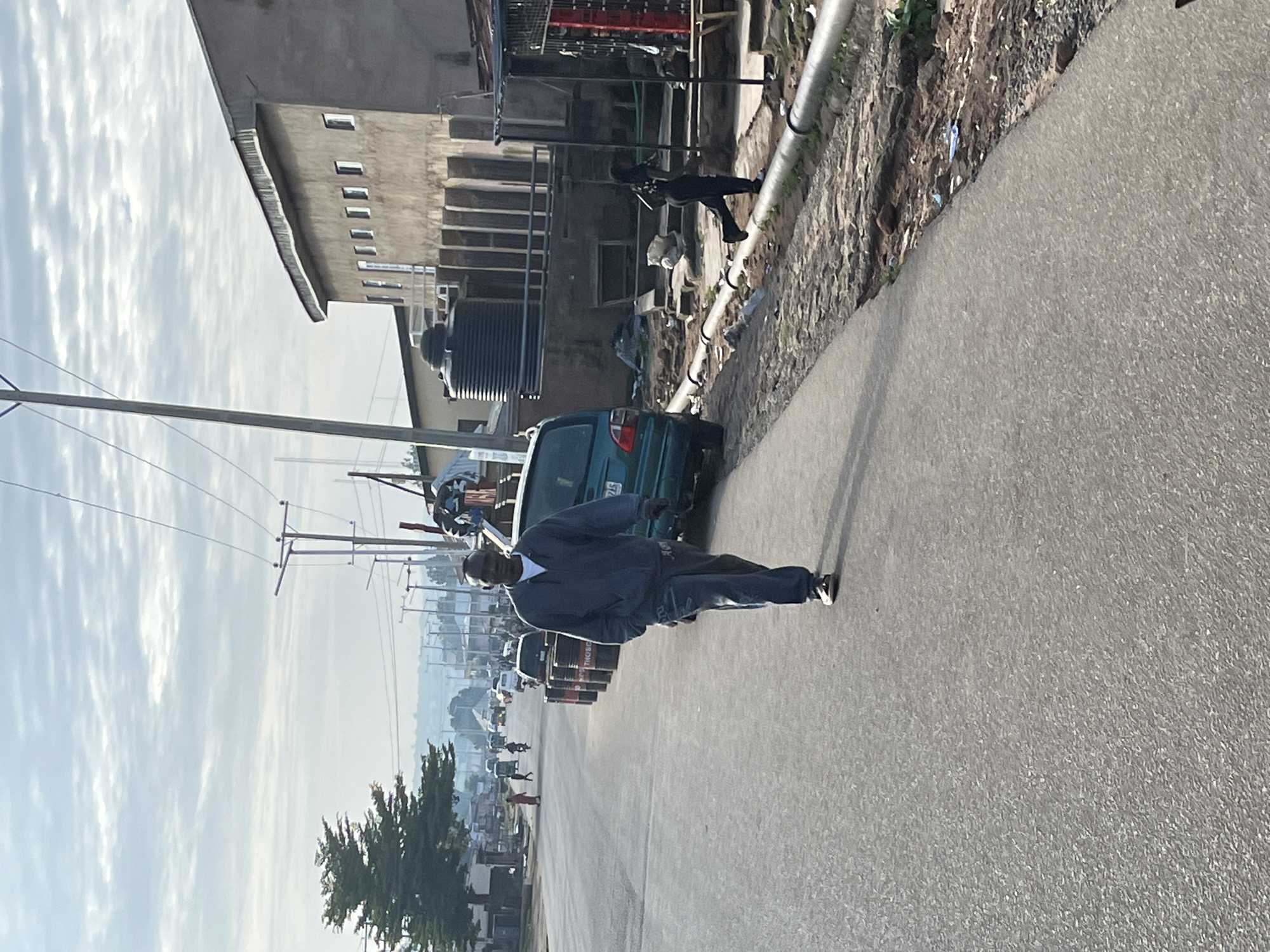 A man is walking on the right side of a paved road in what appears to be an urban environment, possibly in Jos, Nigeria. He is wearing a dark grey hooded jacket over a white t-shirt and dark grey pants. The man is looking towards the camera and walking away from the background. To his right, there is a sidewalk with a rougher surface, and beyond that, a building with multiple windows. A large black water tank is visible near the building. In the foreground, next to the man, are several dark, cylindrical objects, likely barrels, placed in a line along the edge of the road. There is a teal-colored vehicle parked to the right of the barrels. Further down the road, there are other vehicles and people, suggesting ongoing activity. The sky is overcast with diffused light. The overall scene depicts a typical street scene with infrastructure and people going about their day.