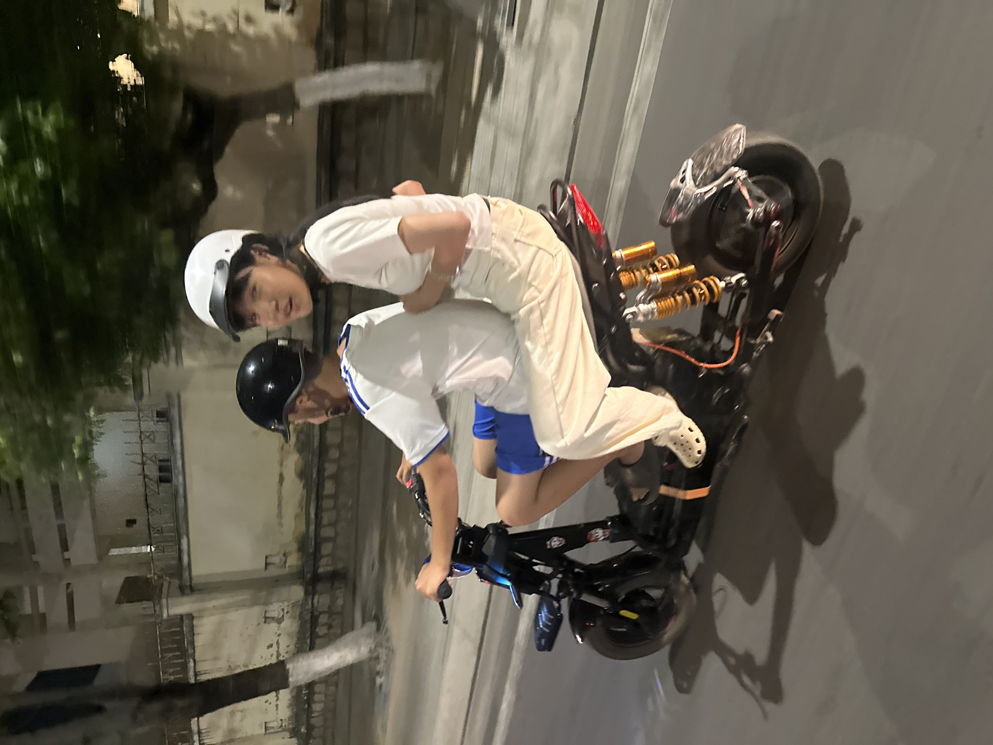 Two people are riding a small motorcycle on a paved road at night in what appears to be the Bac Nha Trang Ward area of Vietnam. The driver wears a black helmet, a white t-shirt with blue stripes on the shoulders, and blue shorts. The passenger wears a white helmet, a white t-shirt, and light-colored pants. They are both leaning forward on the motorcycle. In the background, there are buildings and trees.