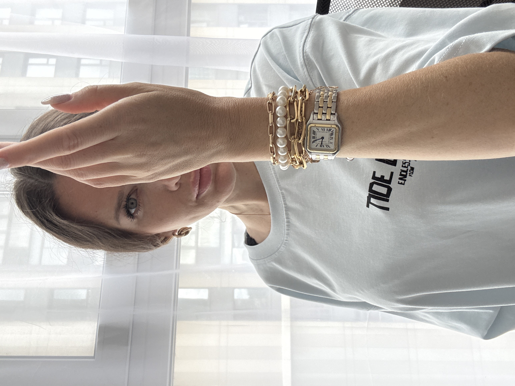 A woman is pictured from the chest up, holding her right hand in front of her face, partially obscuring it. She is wearing a light blue long-sleeved t-shirt with black text on the front that reads "TIDE" and "ENDLESS FLOW". On her left wrist, she has multiple bracelets and a two-tone watch with a silver face and gold accents. She is also wearing gold hoop earrings and a thin gold necklace. The background consists of a window with sheer white curtains. The scene appears to be indoors.