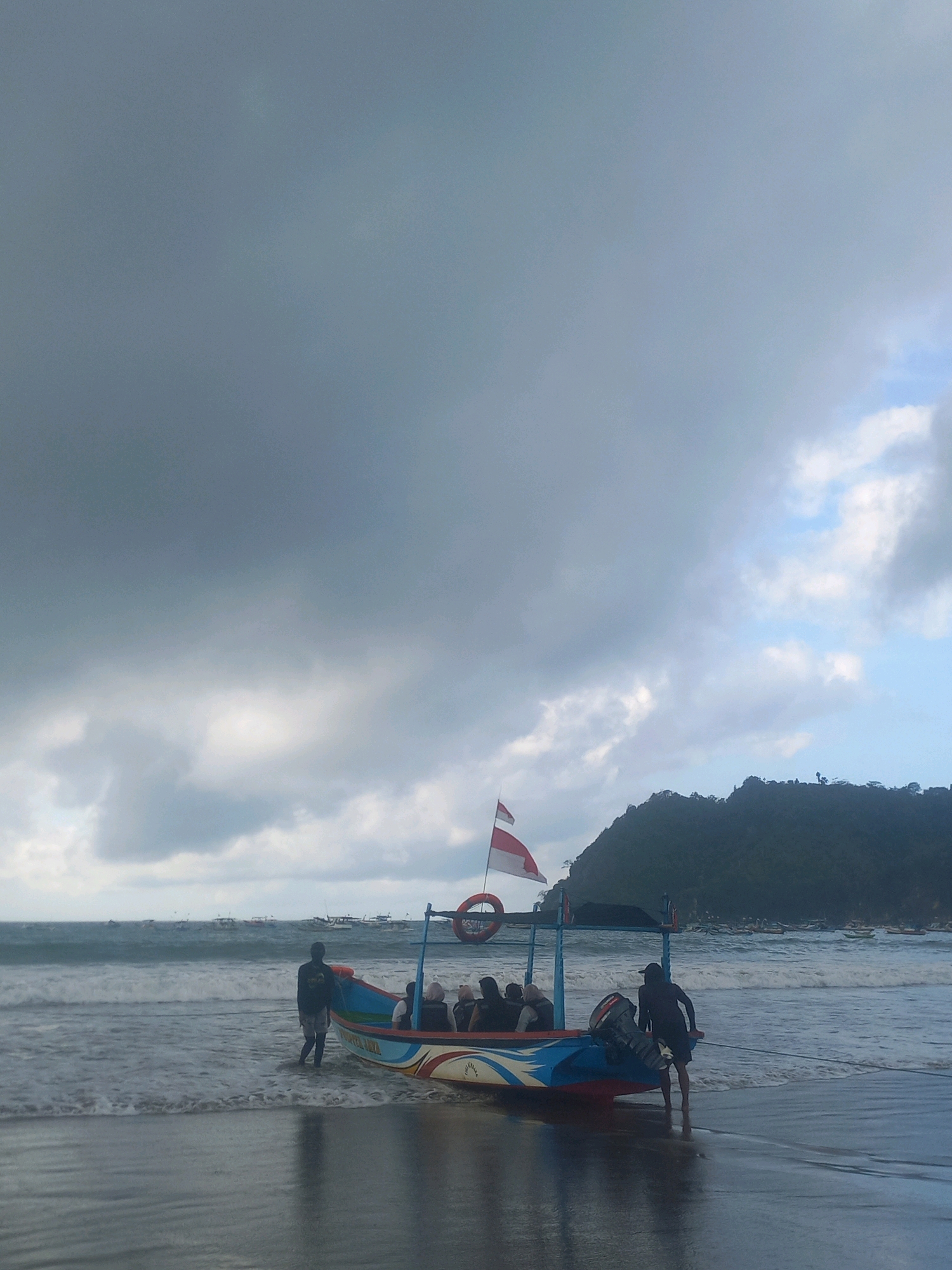On a beach in an unidentified city in Indonesia, two individuals are standing by a colorful fishing boat filled with passengers. The boat, with an Indonesian flag flying from its stern, is partially submerged in the gentle waves. The water is a dark, foamy blue, reflecting the overcast sky. In the background, a lush green mountain rises steeply from the coastline, its peak shrouded in clouds. Further out at sea, a fleet of smaller boats dots the horizon. The beach itself is a dark, wet sand, with the tide reaching the boat and the legs of the two people near it. The overall mood of the image is somewhat somber due to the cloudy weather, but also peaceful due to the tranquil scene.
