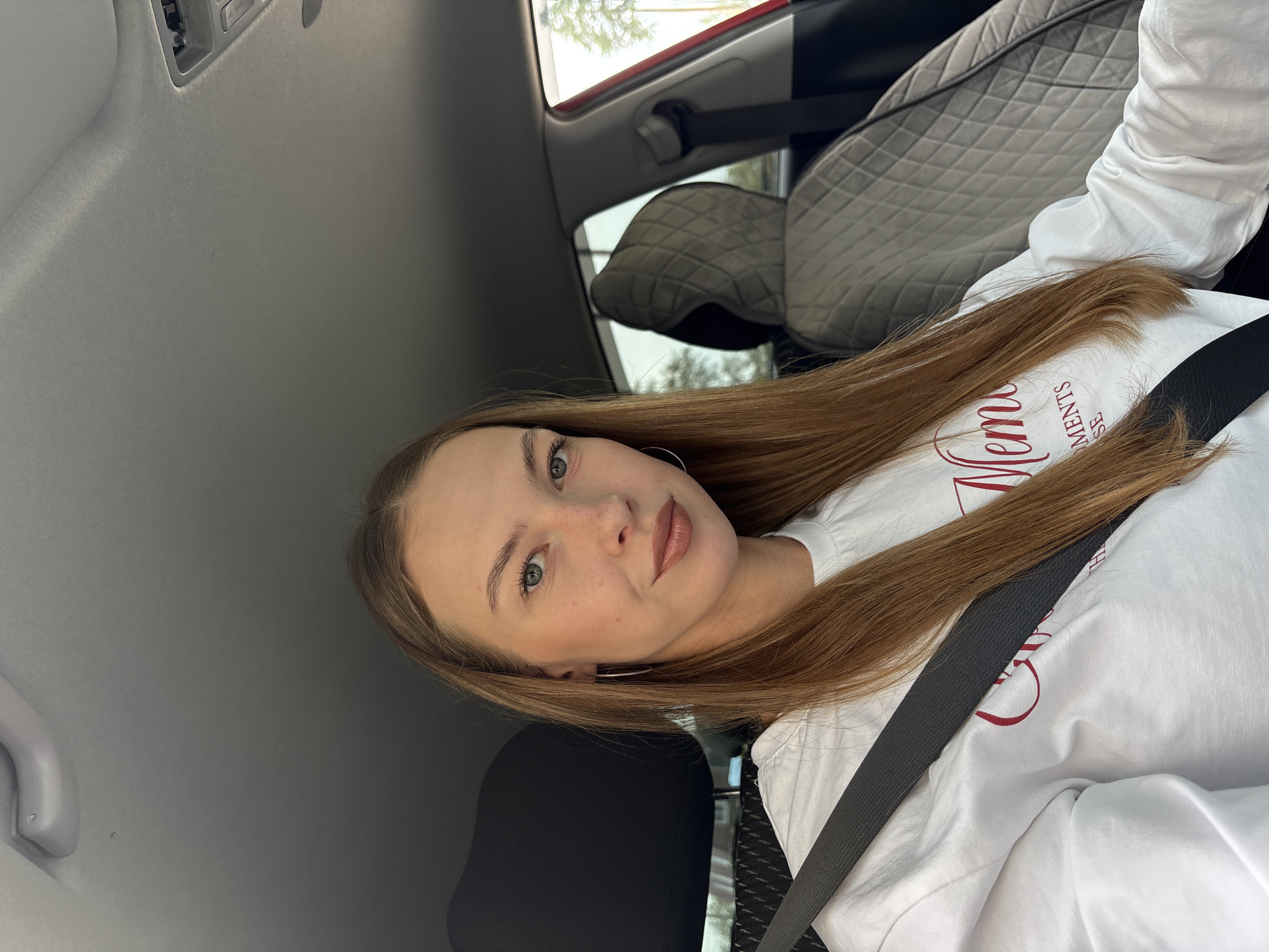 This is a portrait-oriented, selfie-style photo taken from inside a car, likely in Perm, Russia. The subject is a young woman with long, reddish-brown hair, wearing a white long-sleeved shirt. She is looking directly at the camera with a slight smile and her eyes are open. The background shows the interior of the car, including a quilted headrest of a passenger seat and the car's ceiling, with a door handle visible on the left side. The overall lighting suggests it is daytime.
On the woman's shirt, there is visible text in red lettering that reads "Moments". The image is slightly blurry in some areas, particularly around the edges and the text. The focus is on the woman's face and upper body. The photo captures a casual moment, possibly during a car ride. The angle and composition indicate it was taken by the subject herself.