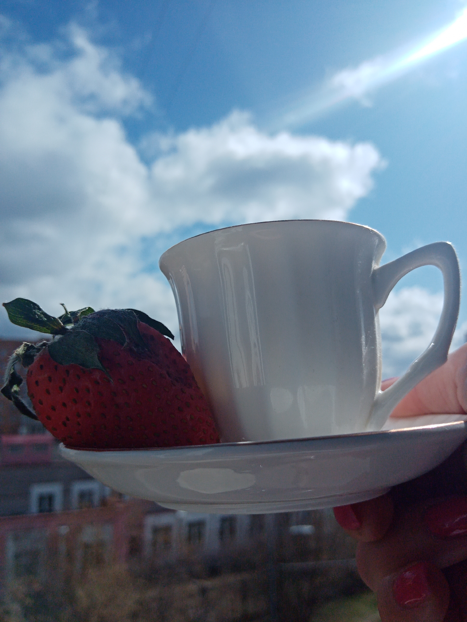 A white teacup and saucer are held in someone's hand in front of a cloudy blue sky. Leaning against the cup is a large strawberry. Buildings are visible in the background. The photograph was taken in Elektrostal, Russia.