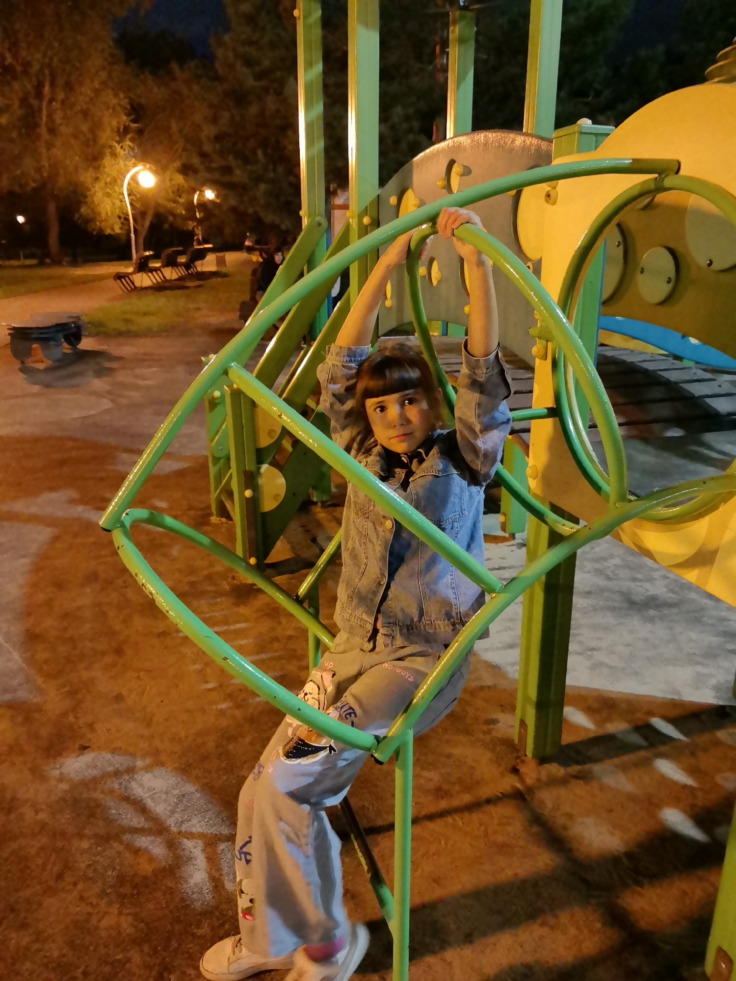 In this nighttime photograph, a young girl is the central subject, posed on a playground structure. She is wearing a denim jacket and light-colored pants adorned with cartoon-like graphics. Her posture suggests she is holding onto the green metal frame of the playground equipment. The girl looks directly at the camera with a neutral expression.
The setting is a playground at night. The area is illuminated by streetlights, casting a warm glow on the scene and creating distinct shadows on the ground. In the background, trees and more playground equipment, primarily in yellow and green, are visible. The overall ambiance is quiet and still, characteristic of a park after dark. There is text on the girl's pants that reads "No boys." The scene appears to be a public park in Kazan, Russia, as indicated by the location context. The weather is not explicitly discernible, but the lighting suggests it is a clear night.