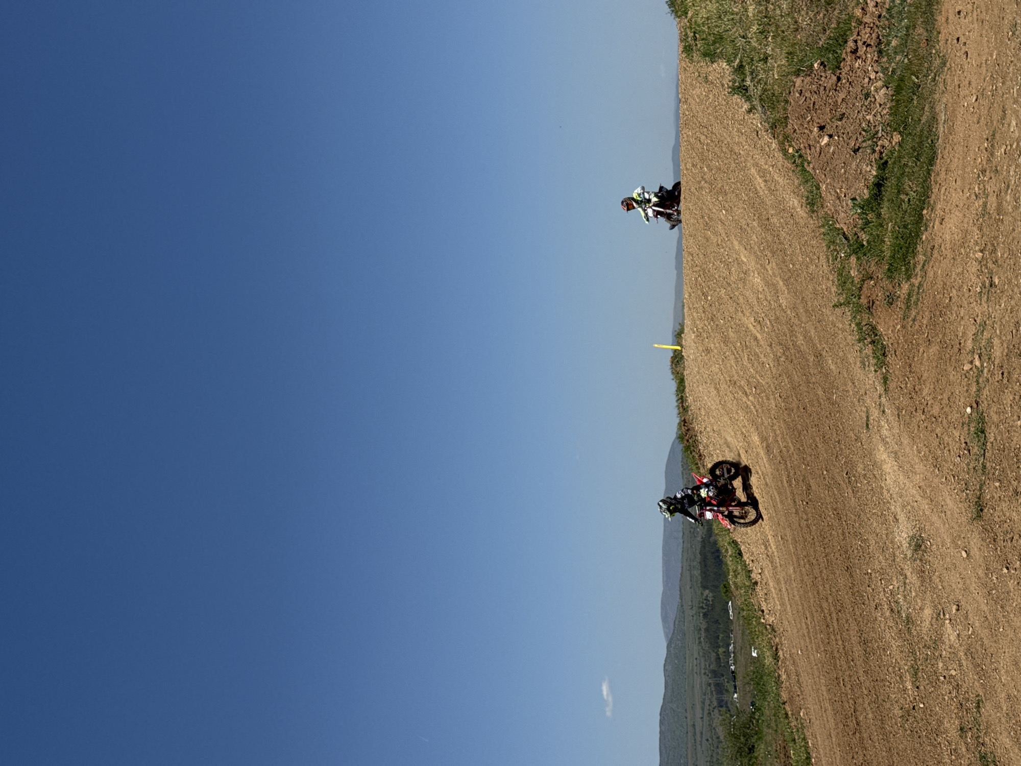 The image shows two dirt bike riders at a motocross event in Selishte, Bulgaria. The sky is clear and blue. One rider is positioned near the top of a dirt track, while the other is closer to the bottom. The track is dirt with some vegetation on the edges. A yellow marker is visible on the track near the top rider. Mountains are visible in the background.