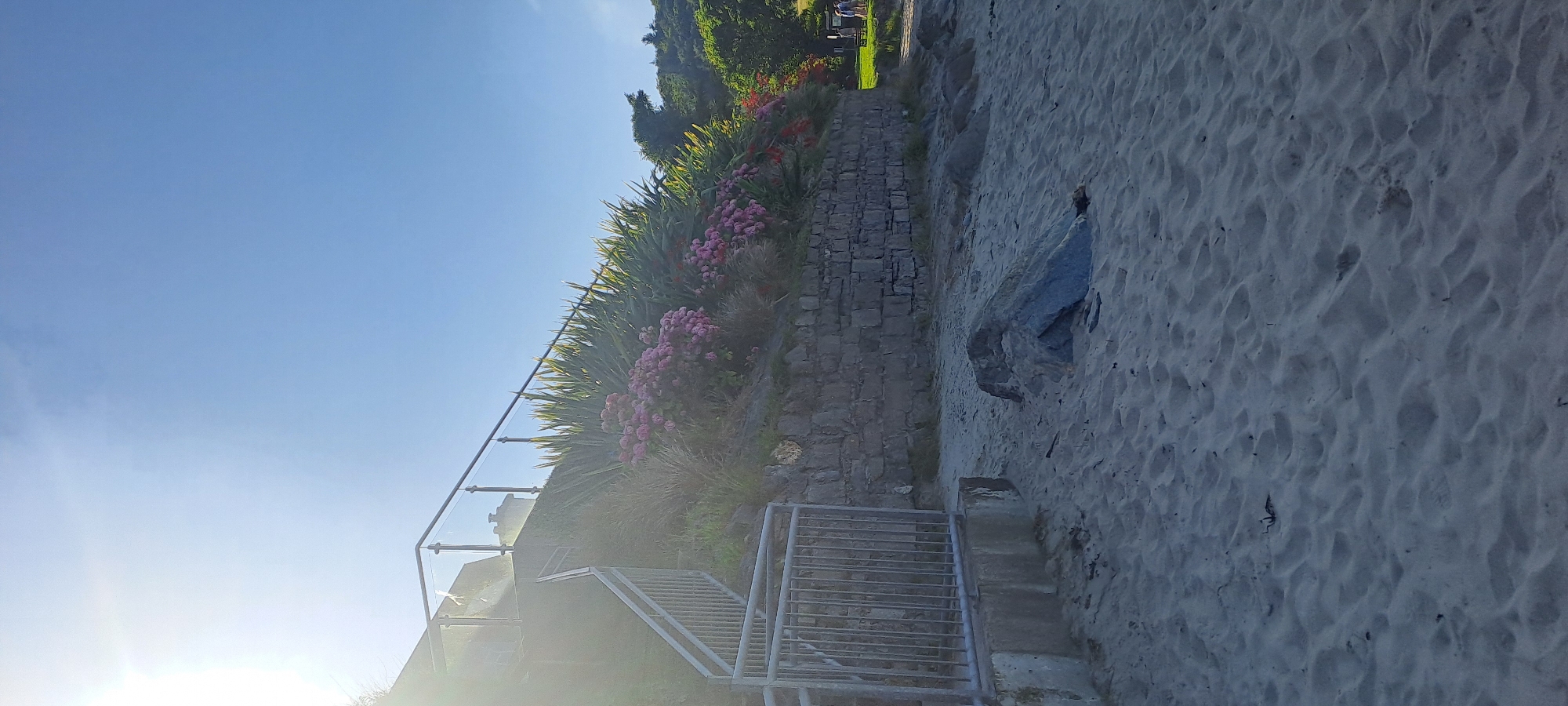 A sun-drenched view of a sandy beach in an unknown location in Ireland, with a stone wall and lush vegetation rising from the sand. A metal and glass railing tops the stone wall, adjacent to a concrete staircase leading upwards. The sand is textured with ripples and a few scattered rocks. In the background, trees and a glimpse of what appears to be a building are visible, along with two individuals walking in the distance. The sky is a clear blue, with the sun creating a lens flare on the left side of the image.