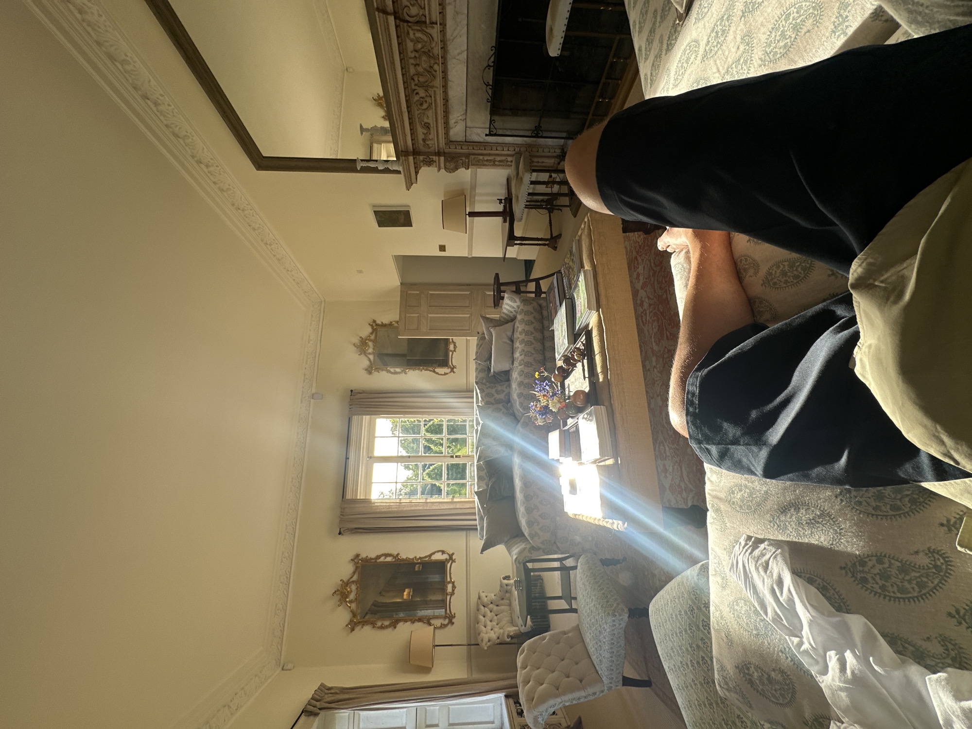 A casually composed interior shot reveals a living room with ornate, classical architectural detailing, including decorative moldings and a fireplace mantel with a large mirror above it. Natural light streams in from a window, illuminating the room and casting reflections on the coffee table and seating areas. A person is partially visible on a patterned sofa, wearing dark clothing and tan pants. The room features a mix of furniture styles, including upholstered chairs, a glass-topped coffee table, and decorative accessories. The scene is undisturbed, capturing a moment of relaxation in an elegant setting. The location is Unknown, Unknown.
