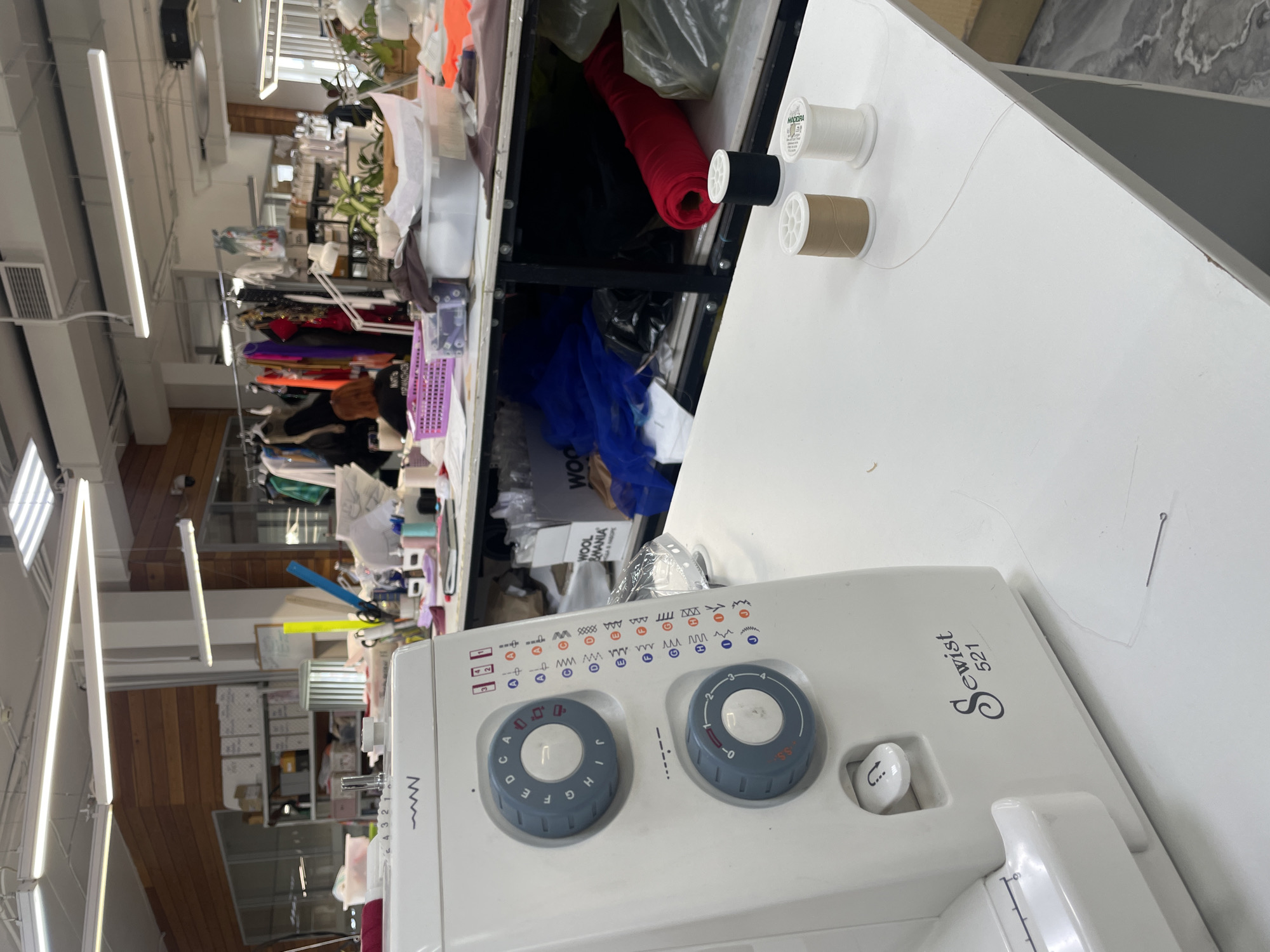 This is a medium-range, eye-level, interior shot of a sewing studio. The studio is well-lit with overhead fluorescent lights. In the foreground, a white sewing machine, identified as a "Sewist 521," occupies the left side of the frame. Its control dials and stitch selection guide are visible. A sewing needle lies on the white tabletop in front of the machine, attached to a thin thread. To the right of the sewing machine are three spools of thread: one black, one beige, and one white.
The middle ground features a shelf unit filled with various items. Notable are rolls of fabric, including red fabric, and stacks of papers, possibly patterns. A person, seen from the back with dark hair, is partially visible at a table further back, wearing a dark shirt and working with a purple basket. A variety of clothing items hang on a rack in the background, suggesting an active workshop. The walls are lined with wooden panels and white shelving units. The floor appears to be a light gray laminate. The scene depicts a functional sewing environment, likely in a commercial or educational setting within Pargolovo, Russia.