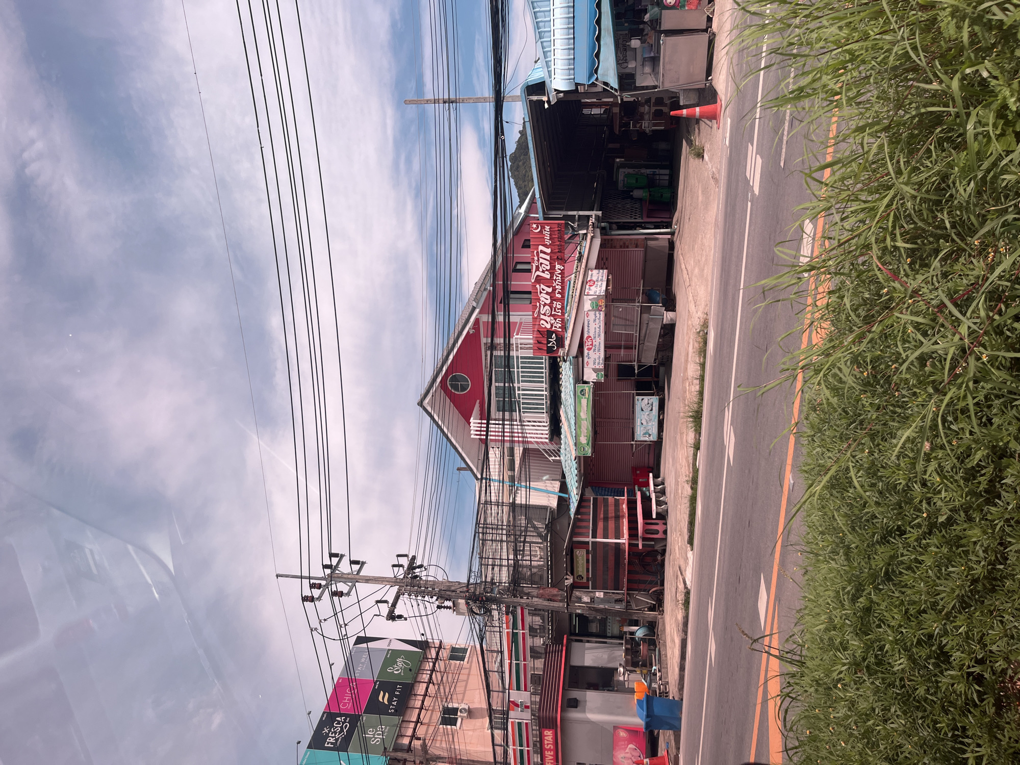 The image shows a street scene in Rawai, Thailand, on a cloudy day. The foreground is dominated by lush green grass and foliage. In the midground, a street runs horizontally, with a yellow line marking the lanes. On the other side of the street, there are several buildings and stalls.
To the left, a pink building with a 7-Eleven sign is visible. Next to it is a smaller structure labeled "FIVE STAR". A blue trash bin and an orange traffic cone are placed near the curb.
In the center, a bright pink building with white balconies stands out. This building has a sign with Thai script and a "Halal" symbol. In front of it are several small food stalls with striped awnings and open serving areas.
Further to the right, there are more food stalls and shops with various awnings and structures, some with blue roofing. A tall wooden pole with electrical wires extends upwards. In the background, a lush green mountainside is visible under the cloudy sky. The overall scene depicts a bustling commercial area with a mix of shops, food stalls, and utility infrastructure.
