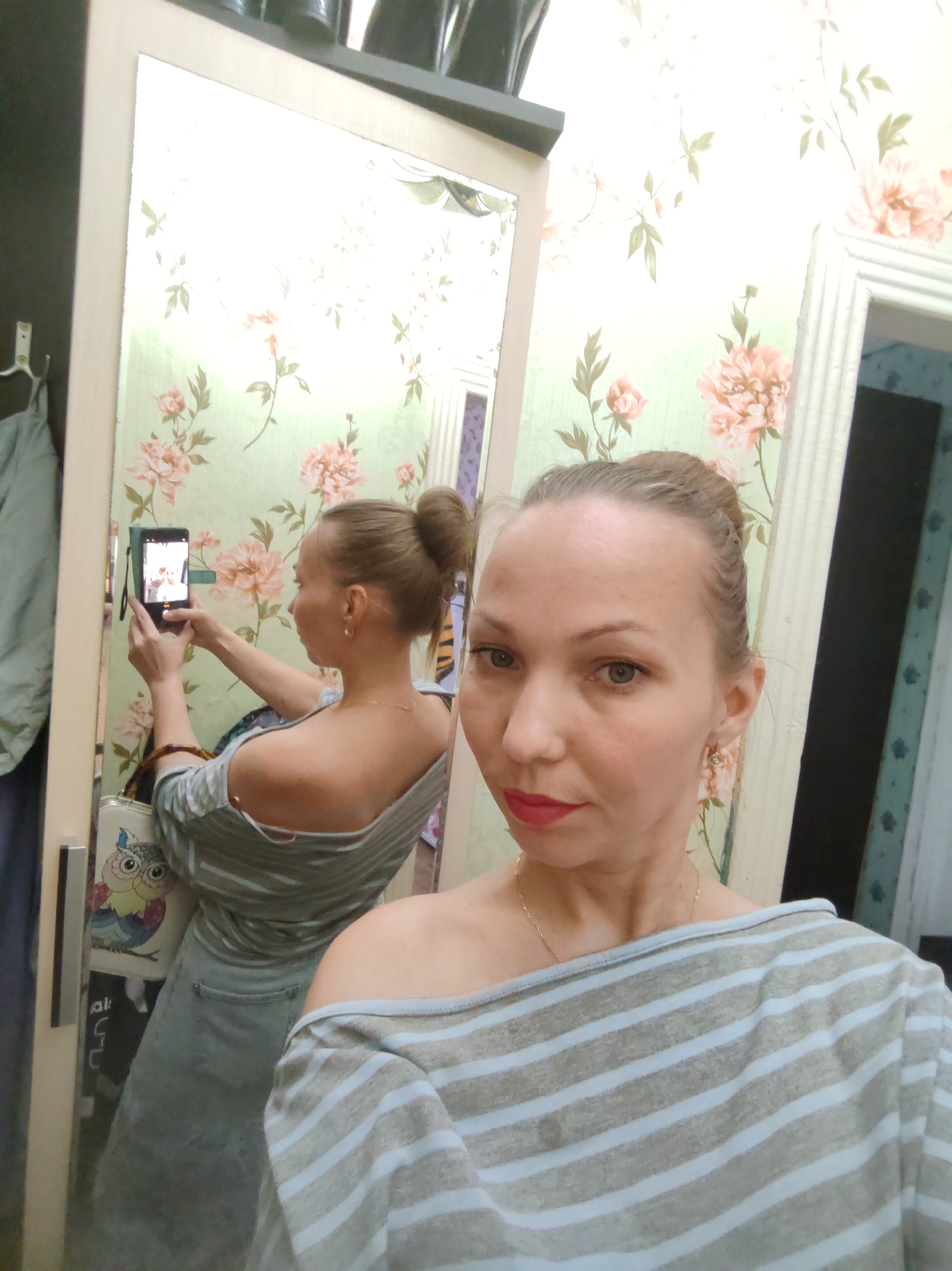 A woman is taking a selfie in front of a mirror. The mirror is mounted on a cabinet in a room with floral wallpaper. The location is identified as Чухлэм, Russia. The woman is wearing a blue and grey striped off-the-shoulder top and has her hair in a bun. Her reflection in the mirror shows her holding a phone to take the picture. A handbag with an owl design is visible in the reflection. A doorway is visible in the background, leading to another room.