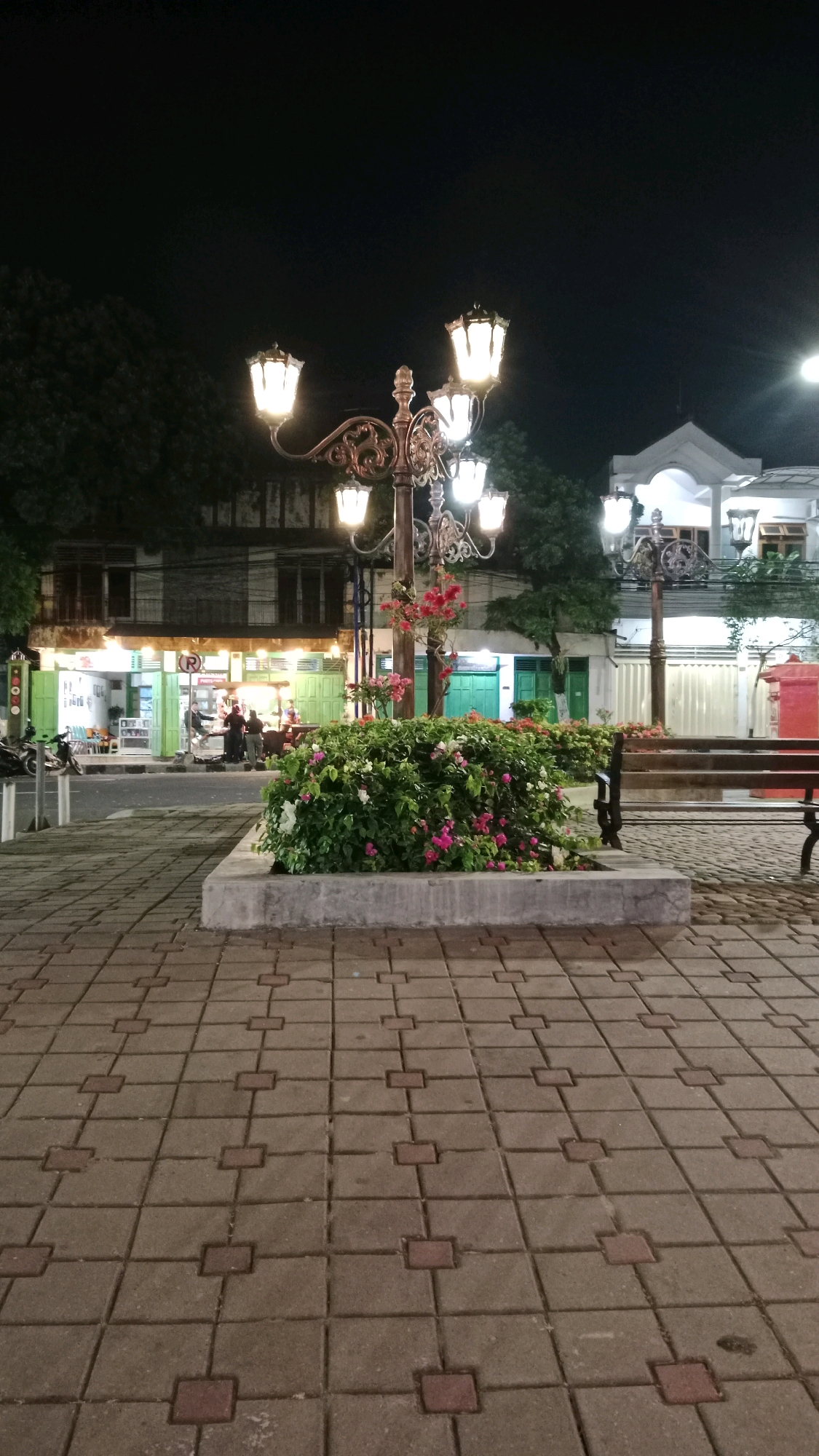 The image depicts a street scene at night in Magetan, Indonesia, likely near a market or commercial area. In the foreground, a decorative lamppost with multiple illuminated bulbs stands beside a well-maintained flowerbed filled with greenery and pink and white flowers. The ground is paved with interlocking square tiles.
Behind the flowerbed, a row of shops with green shutters and illuminated interiors line the street. Several people are visible in front of the shops, appearing to be engaged in activity, possibly selling or buying goods. Motorcycles are parked to the left of the frame. Further down the street, a building with white accents and balconies is visible, illuminated by lights. A wooden bench is situated to the right, providing a place to sit. The sky above is dark, indicating nighttime. The overall impression is of a lively urban environment after dark.