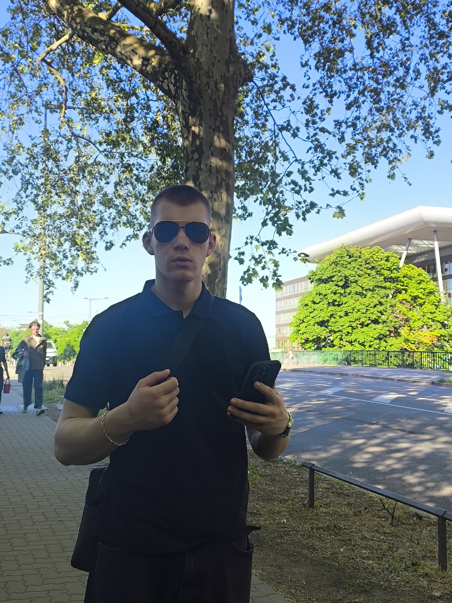 A man stands on a sidewalk beside a tree, holding a smartphone. He is wearing sunglasses, a black polo shirt, and a silver bracelet. He is carrying a black bag across his chest. Two people walk in the background on the sidewalk. A green bush and a road separate the sidewalk from a large building in the background, with a white roof. The location is Strasbourg, France, specifically in front of the European Court of Human Rights.