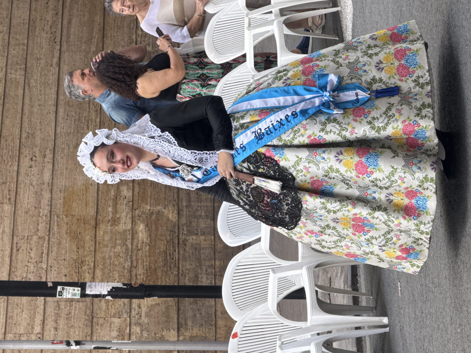 In an outdoor urban setting with a stone wall in the background, a young woman dressed in traditional Valencian attire stands. She wears a voluminous skirt with a floral pattern in shades of pink, blue, and yellow, a black bodice, and an elaborate white lace headdress. A blue and white sash, inscribed with white lettering, is draped across her chest. She holds a fan in her left hand. Several white plastic chairs are arranged behind her, and other individuals, including a man and two women, are visible in the background. The ground appears to be asphalt.